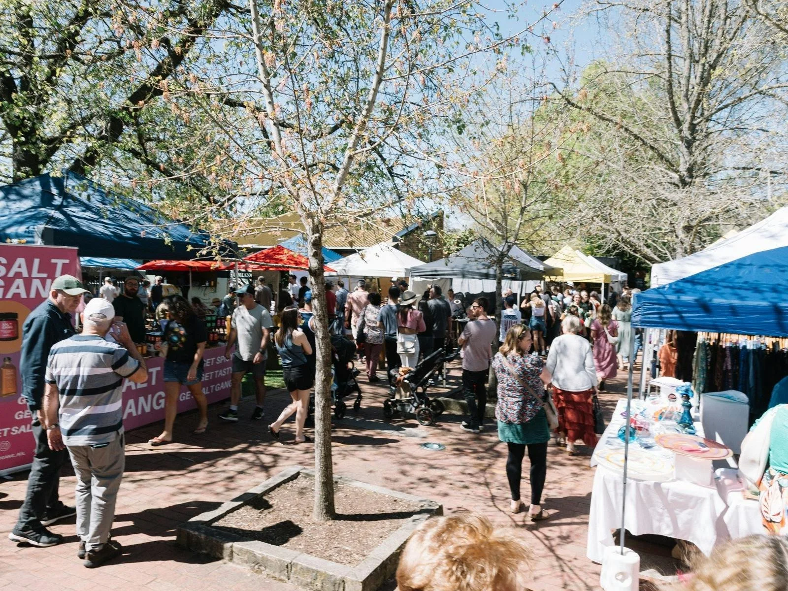 Indoor marketplace with various booths selling crafts and products, people browsing, shopping, and sitting at tables in a large, open industrial-style space with high ceilings and hanging lights.