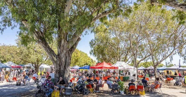 Crowd of people shopping at an indoor market or event, with vendors and sale signs, under a high, glass-paneled ceiling.