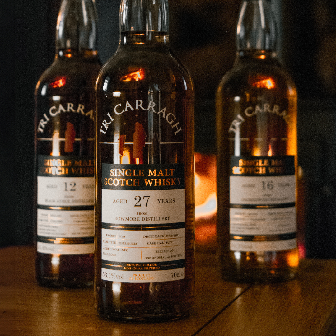 Three bottles of Trig Carragh single malt Scotch whisky, with one in focus showing an age of 27 years, placed on a wooden surface in front of a fireplace.
