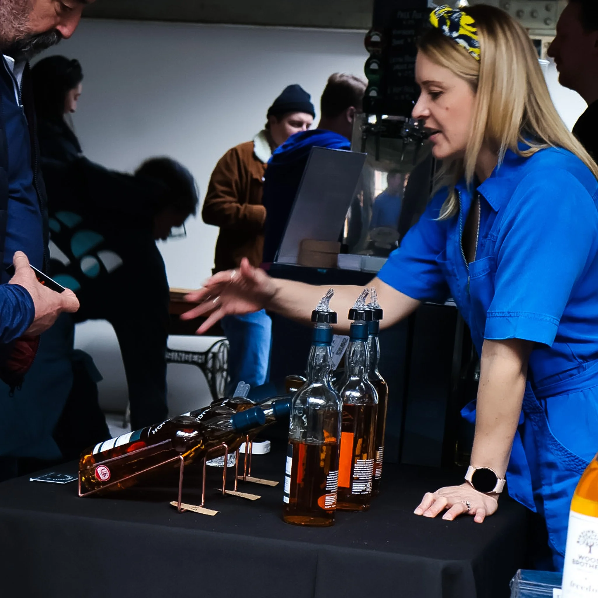 A woman in a blue dress showing bottles of bourbon at a tasting event, with people in the background browsing and talking.