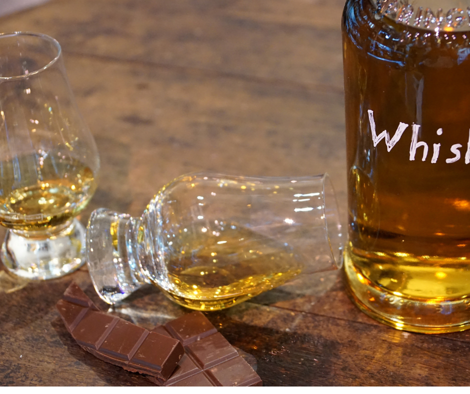 A fallen shot glass and a large bottle of whiskey on a wooden surface, with pieces of chocolate nearby.