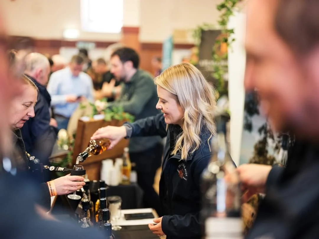 A woman in a black jacket pours beer from a tap into a glass at a crowded indoor event.