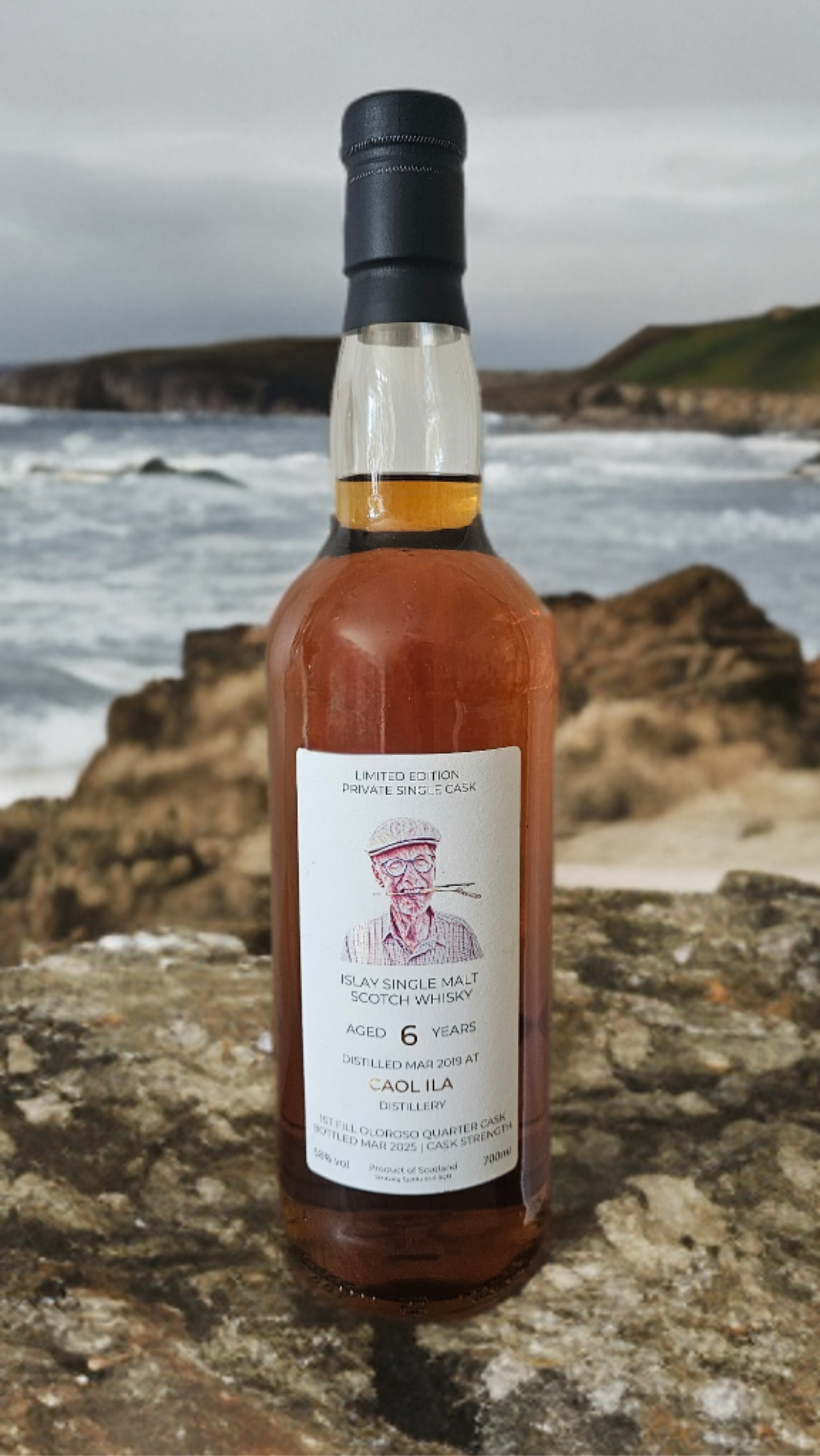 A bottle of Islay single malt Scotch whisky with a white label, black cap, and a small illustration of an elderly man smoking a pipe, placed on a rocky surface with a coastal background of rough sea, rocks, and cliffs.