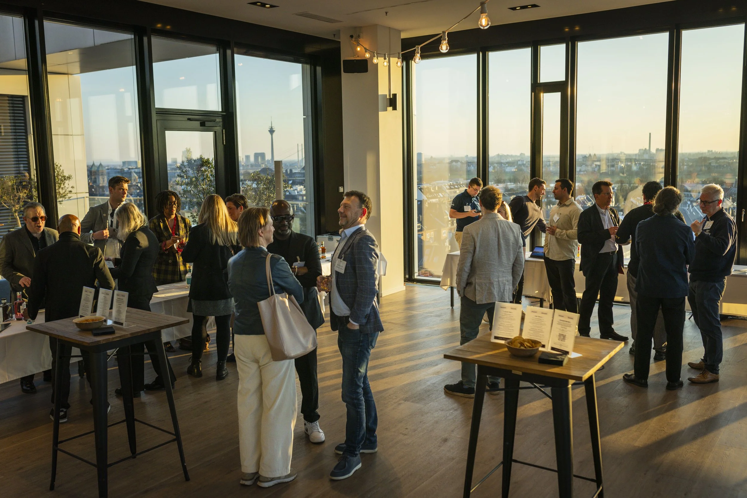 People socializing at a networking event in a modern high-rise venue with large windows and city skyline views during sunset.