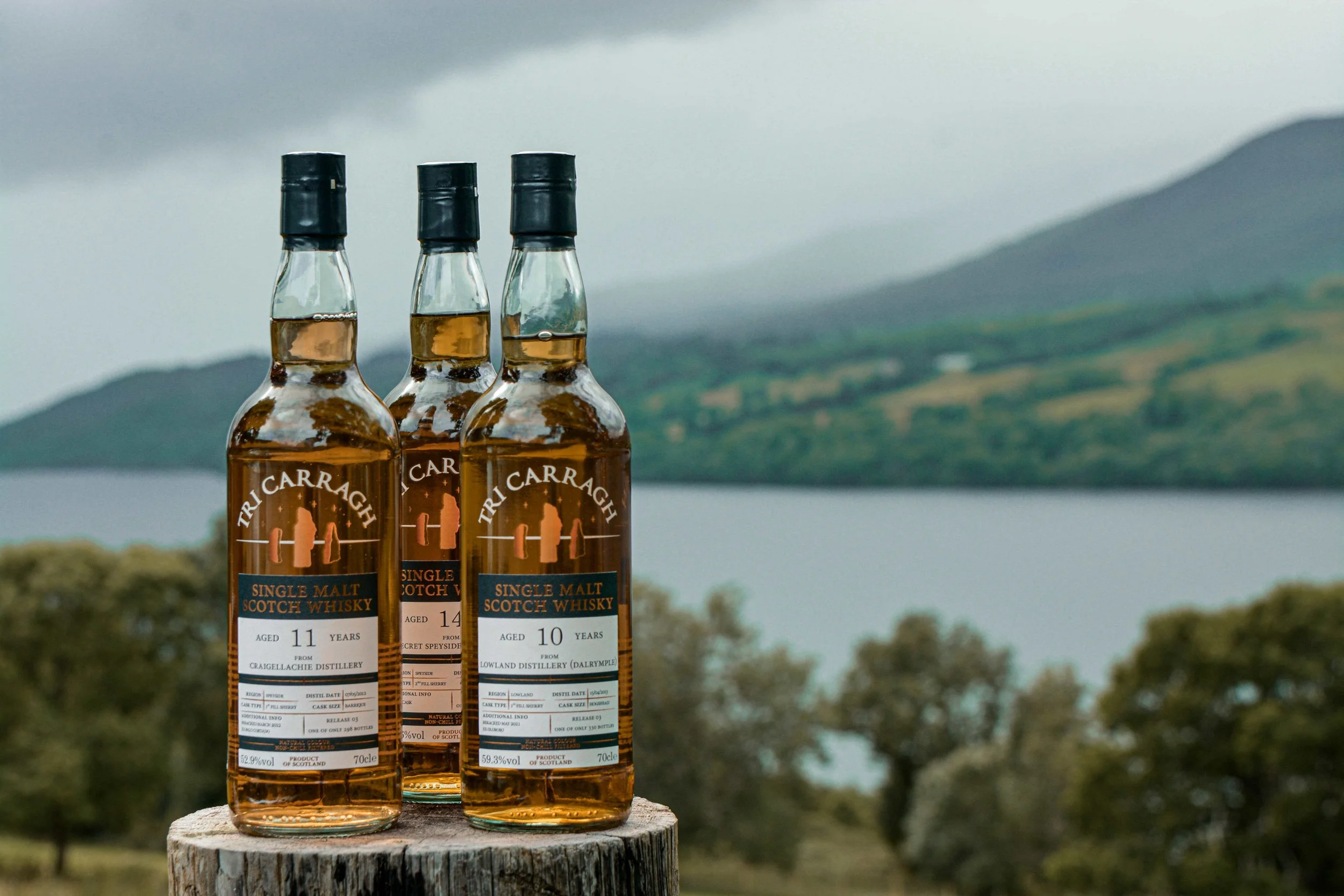 Three bottles of Tricarragh single malt Scotch whisky on a tree stump with a lake, hills, and cloudy sky in the background.