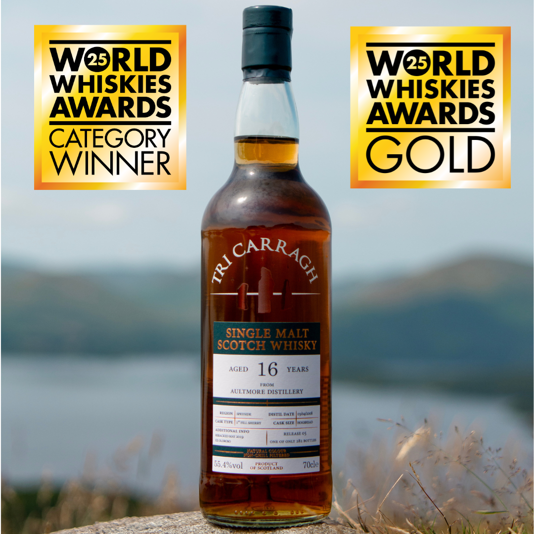 A bottle of Tri Carragh single Malt Scotch whisky with two World Whiskies Awards badges, one for category winner and one for gold. The background shows a landscape with water and hills.
