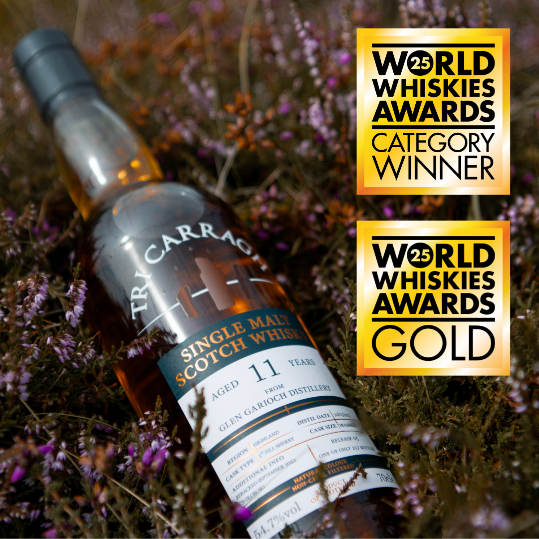 A bottle of Trivh Carrag Scotch Whisky lying among purple flowers with award labels indicating it won gold and category at the 25th World Whiskies Awards.