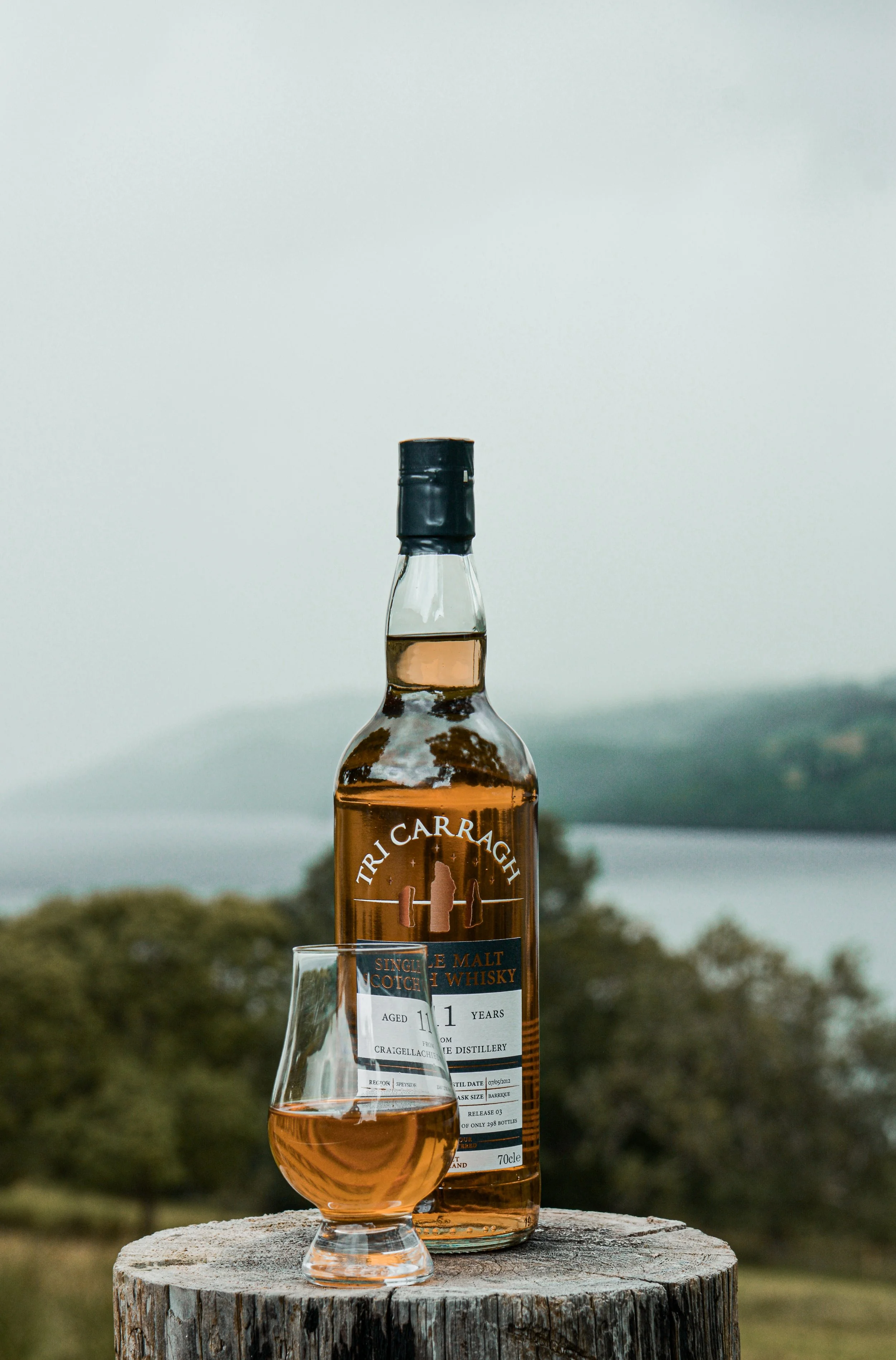 A bottle of Tricarragh single malt Scotch whisky and a glass of whisky on a tree stump with a scenic outdoor background of water and hills.