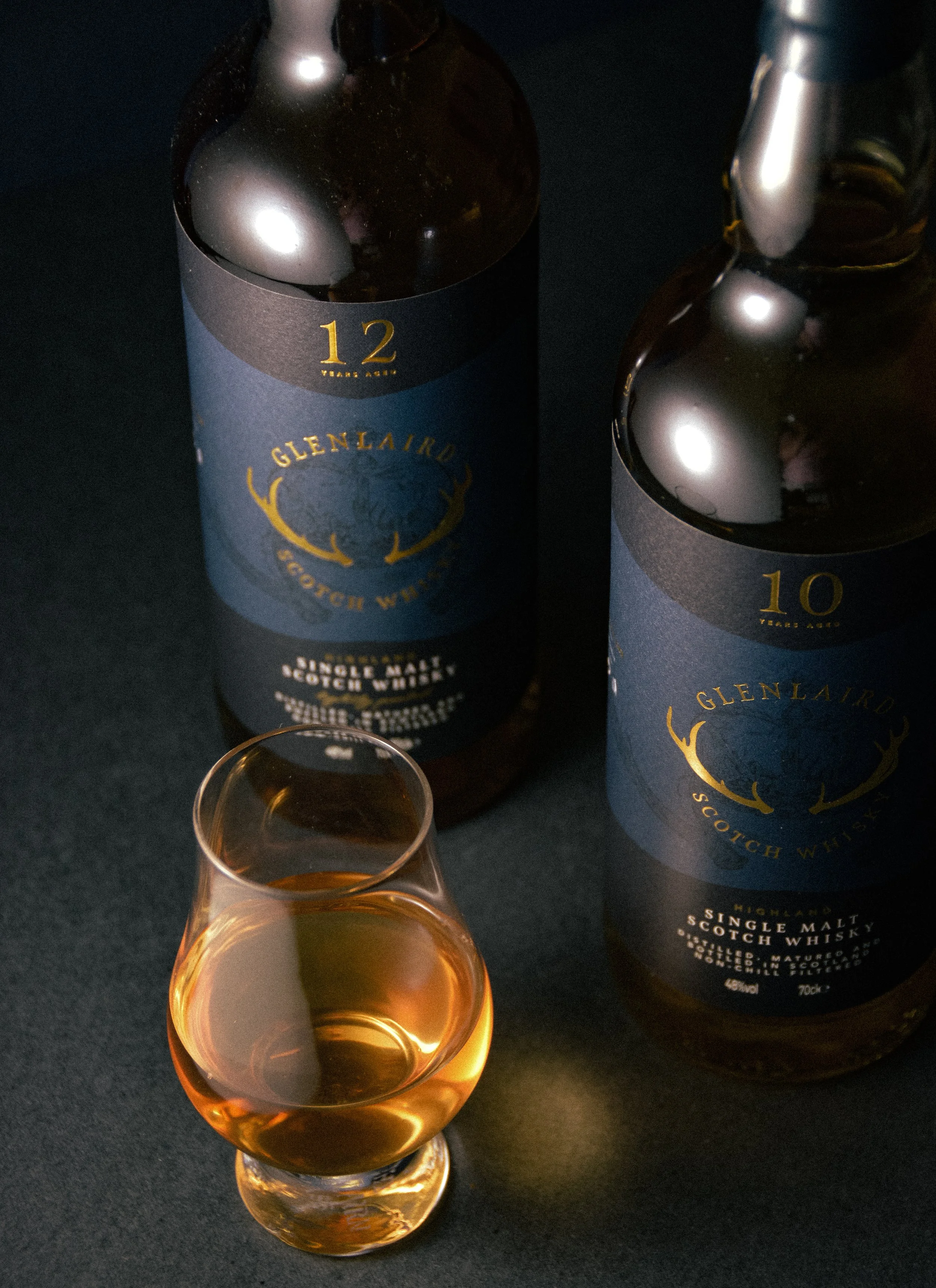 Two bottles of GlenLaird Scotch whisky, aged 10 and 12 years, with a glass of golden whisky in front on a dark surface.