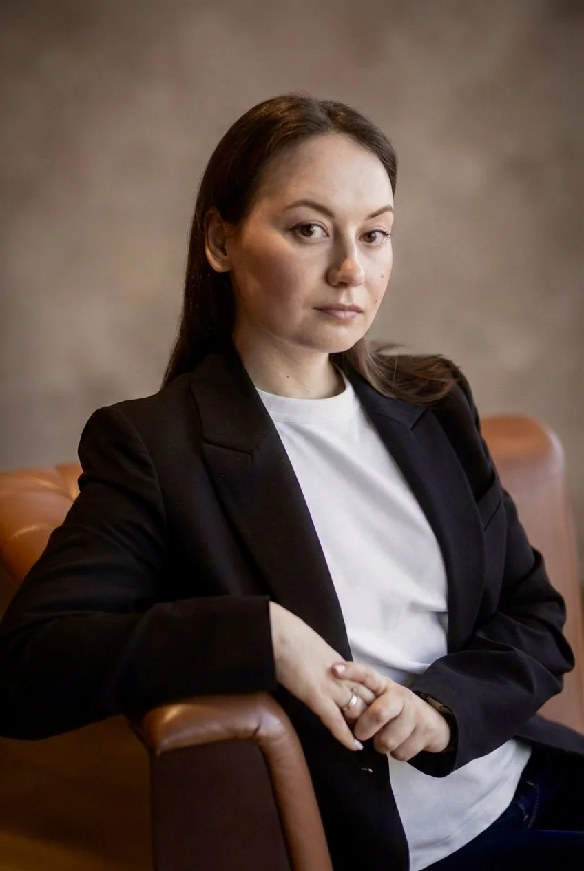 A young woman with brown hair and fair skin, wearing a black blazer and white t-shirt, sitting on a brown leather chair, looking directly at the camera with a neutral expression.
