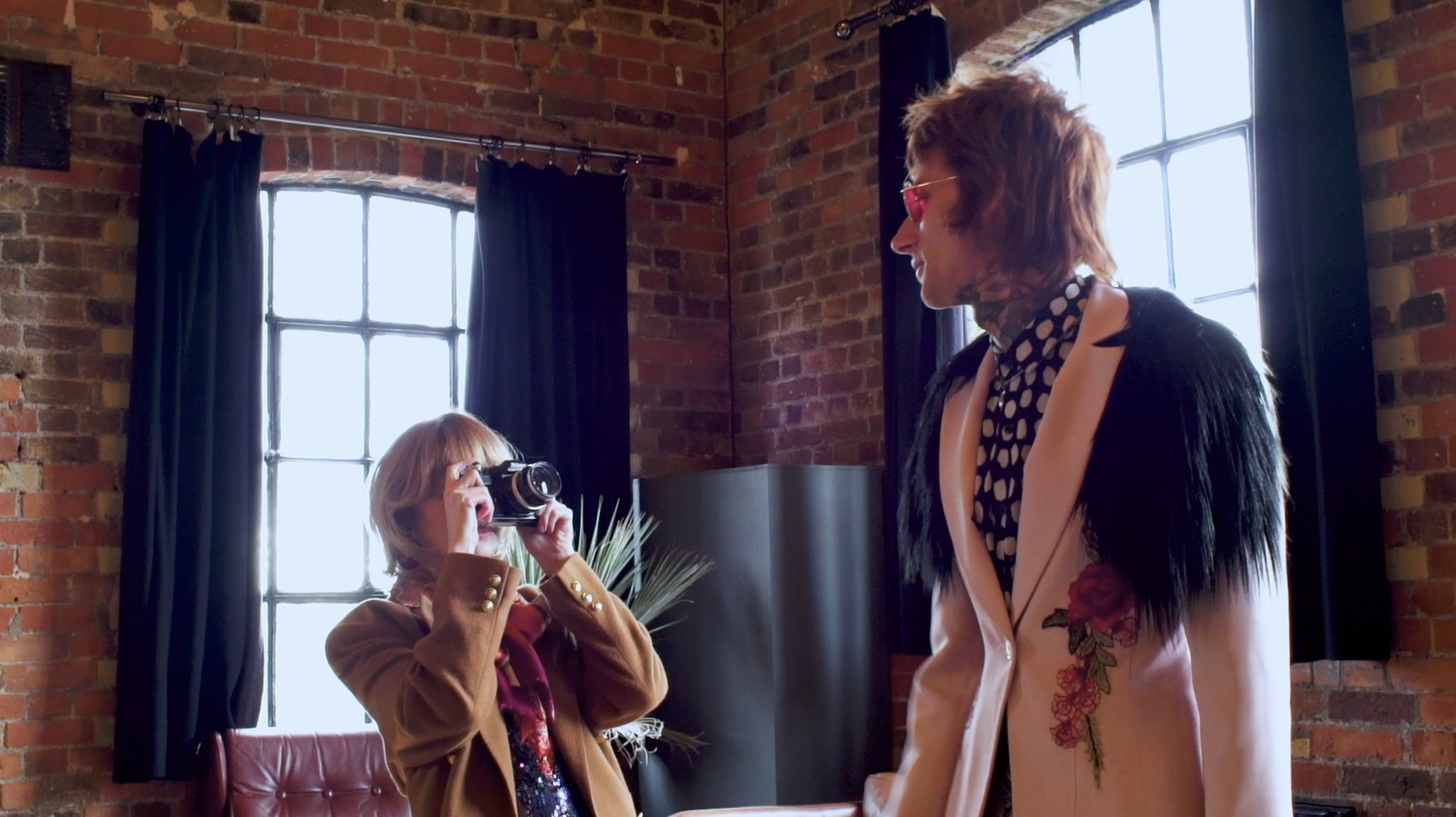 A woman with short hair and sunglasses takes a photo of a man with medium hair and tattoos at a table in a room with brick walls and large windows.