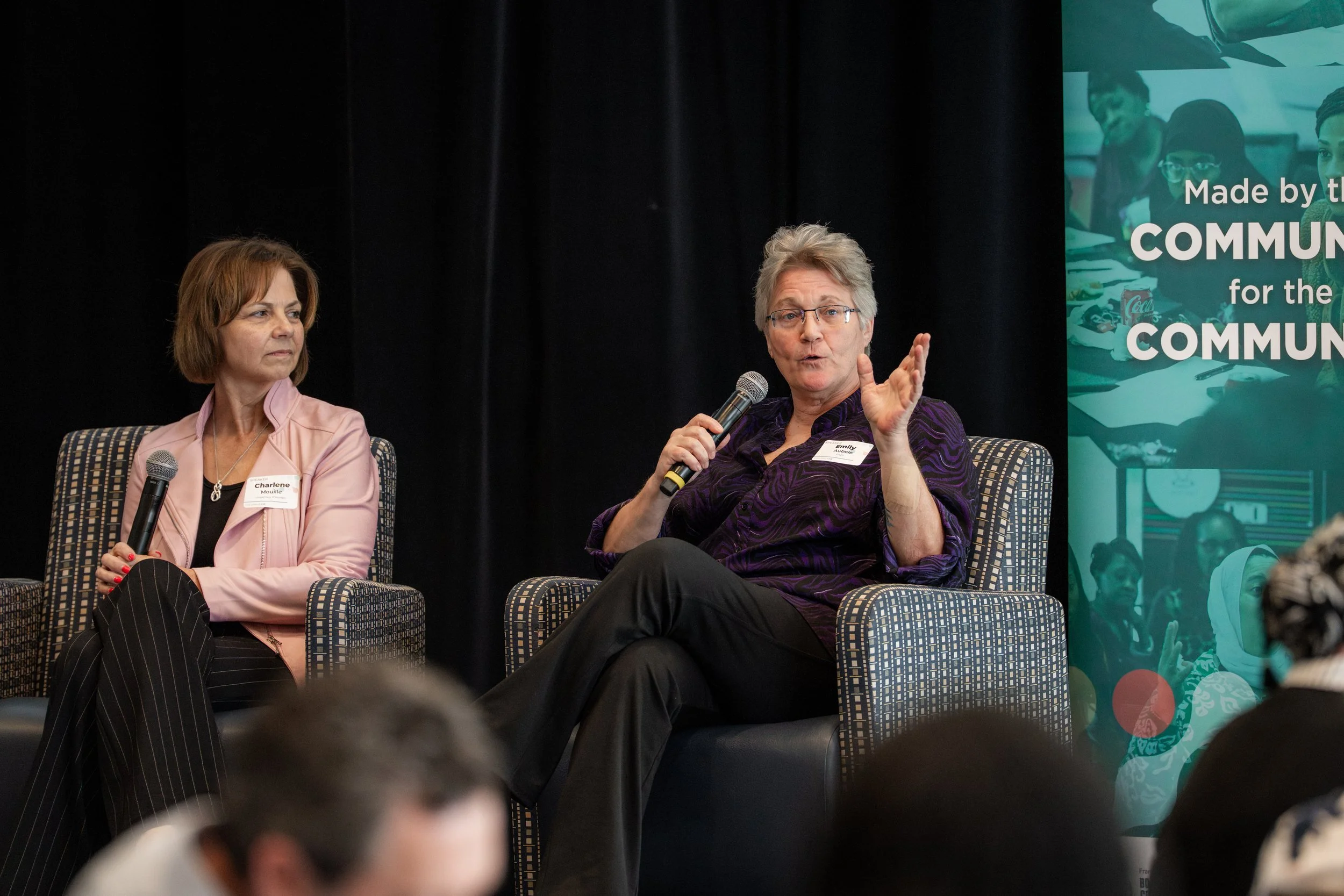 Close-up shot on Charlene Mouille and Emily Aubele sitting in arm chairs on stage, holding microphones. .