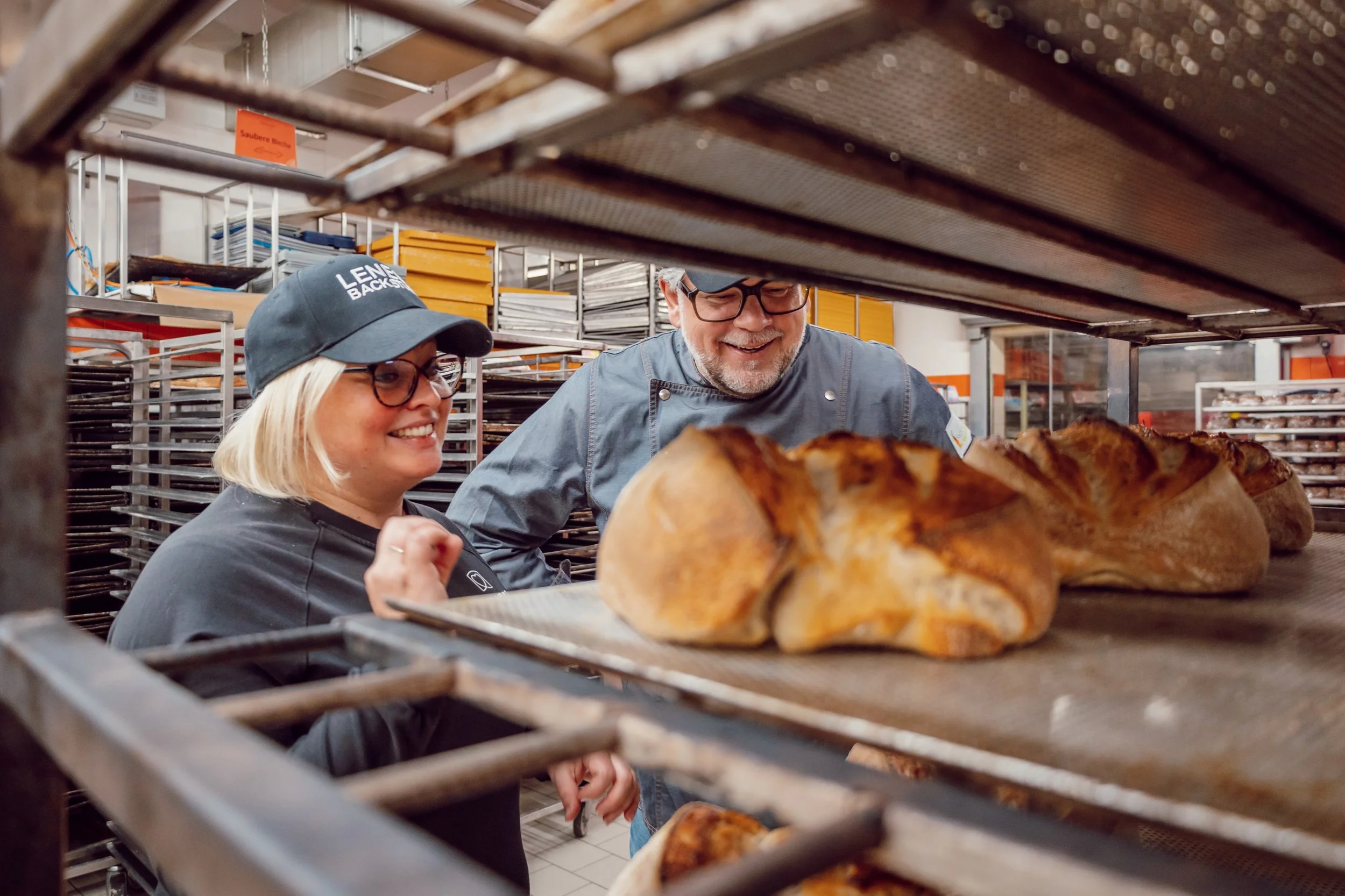 Lene Knoll zeigt André Adden das frisch gebackene Bio Brot. Das gold gebackene Brot liegt in einem Alu-Regal.