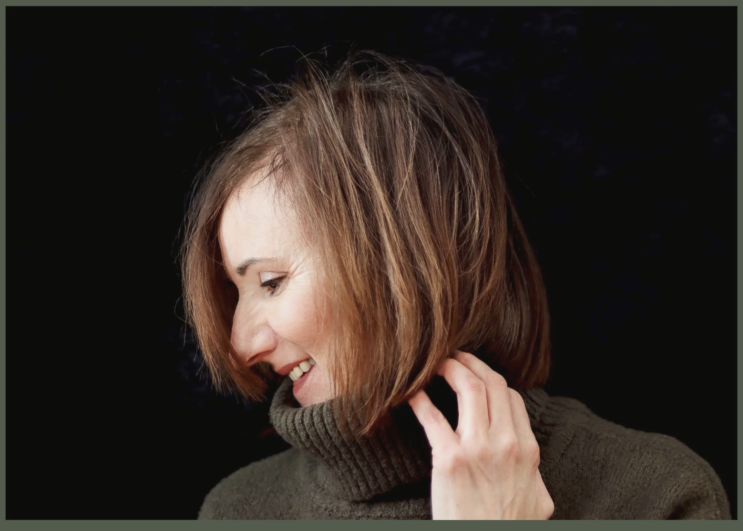 A woman with reddish-brown hair in a bob haircut smiling and touching her neck, wearing a dark turtleneck sweater, against a black background.