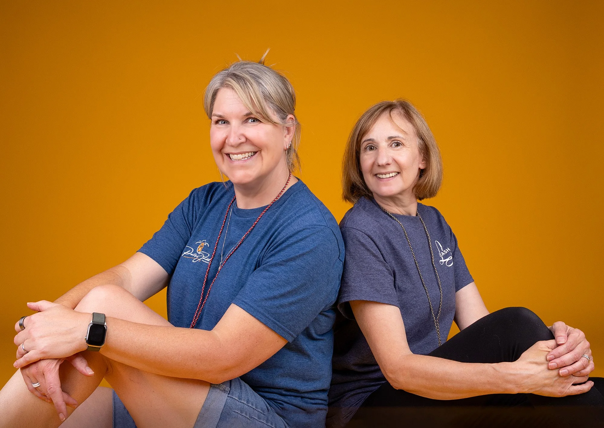 Two women sitting back to back against a yellow background, smiling at the camera, both wearing dark blue shirts and necklaces, with one wearing a smartwatch.