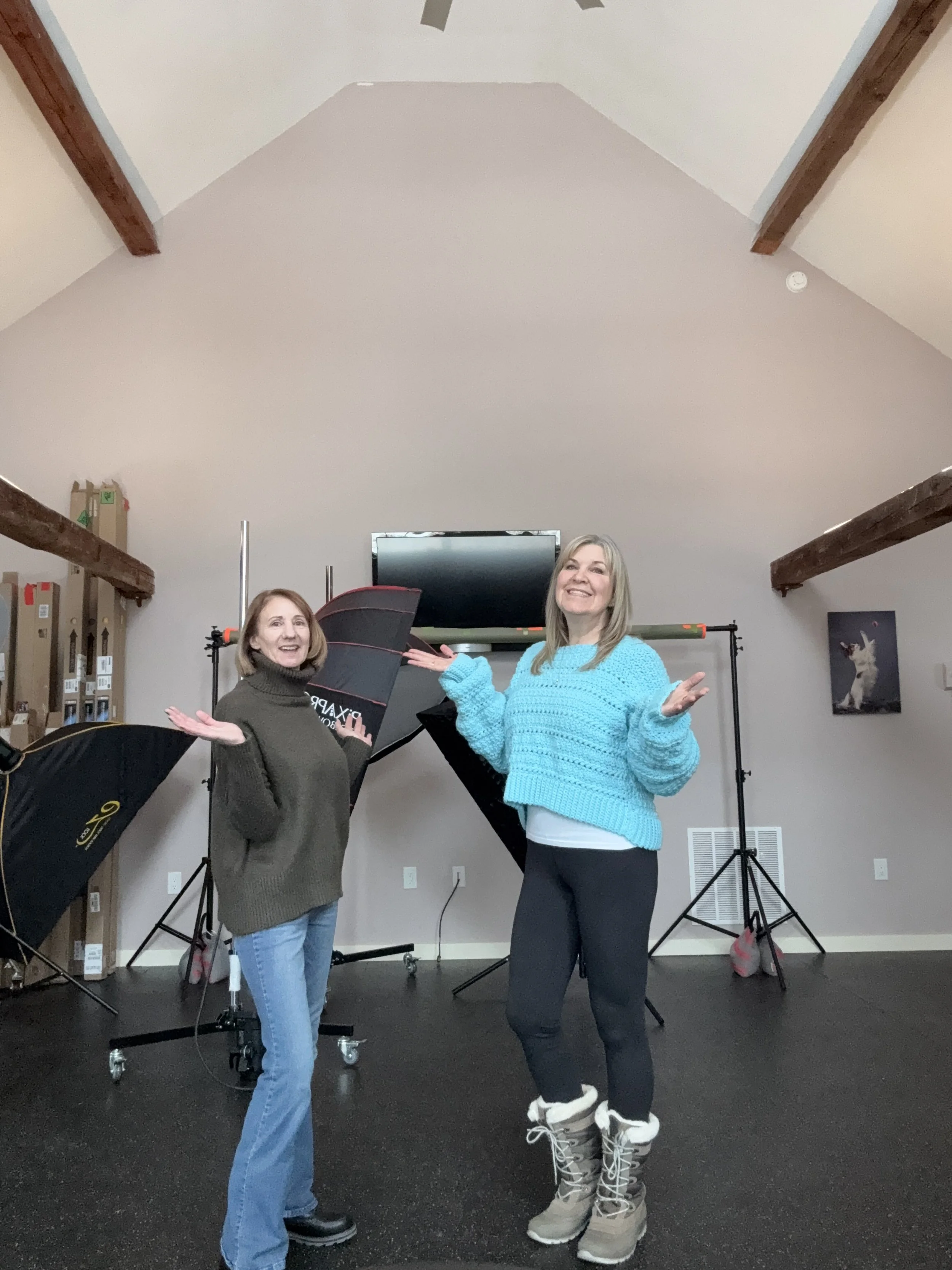 Puptown Girl photographers smiling and posing in a photography studio with professional lighting and a large monitor behind them. One is wearing a gray sweater and jeans, the other a bright blue sweater and winter boots.