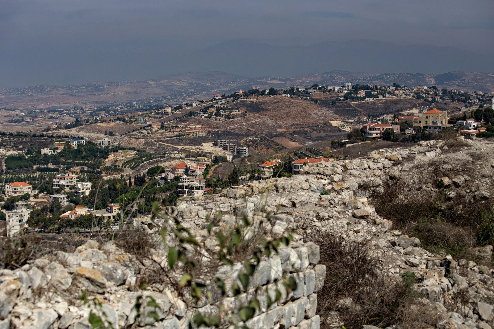 A panoramic view of a hilly landscape with scattered houses, some with orange roofs, and a dry, rocky foreground with sparse vegetation. In the distance, there are more hills and a cloudy sky.