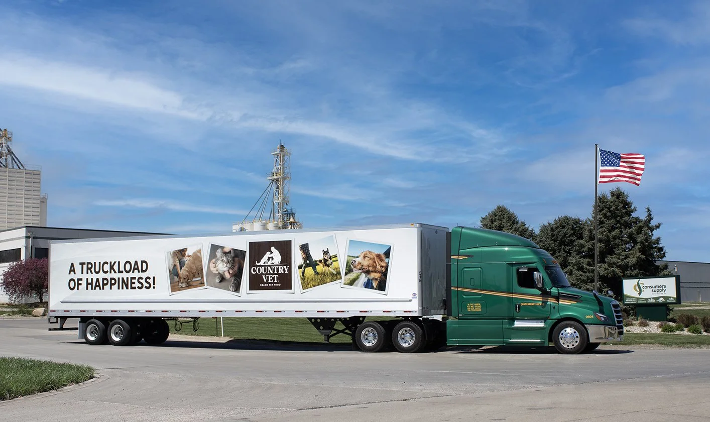 A green semi-truck with a trailer that has images of pets and the words 'A TRUCKLOAD OF HAPPINESS!' and 'Country Vet' on it, parked on a street with an American flag flying nearby under a blue sky.
