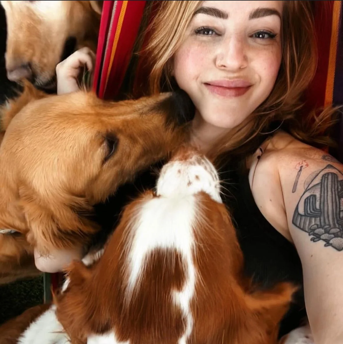 A woman with red hair taking a selfie with three puppies. The puppies are licking her face and appear to be two golden retrievers and a Cavalier King Charles spaniel.