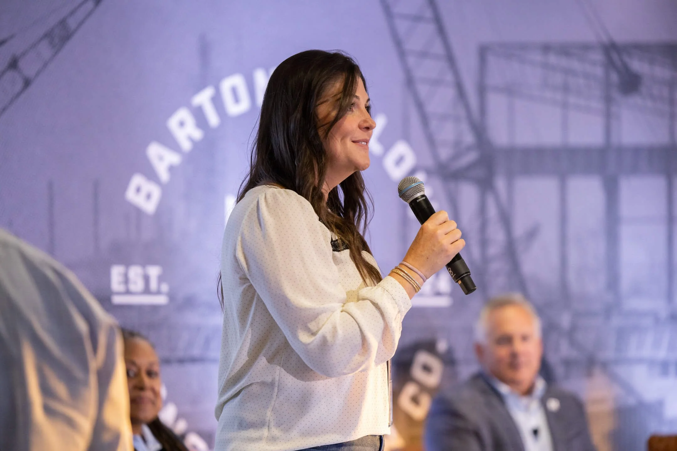 Woman speaking into a microphone at a panel event with a blue background displaying the text "BARTON" and a faint industrial structure.