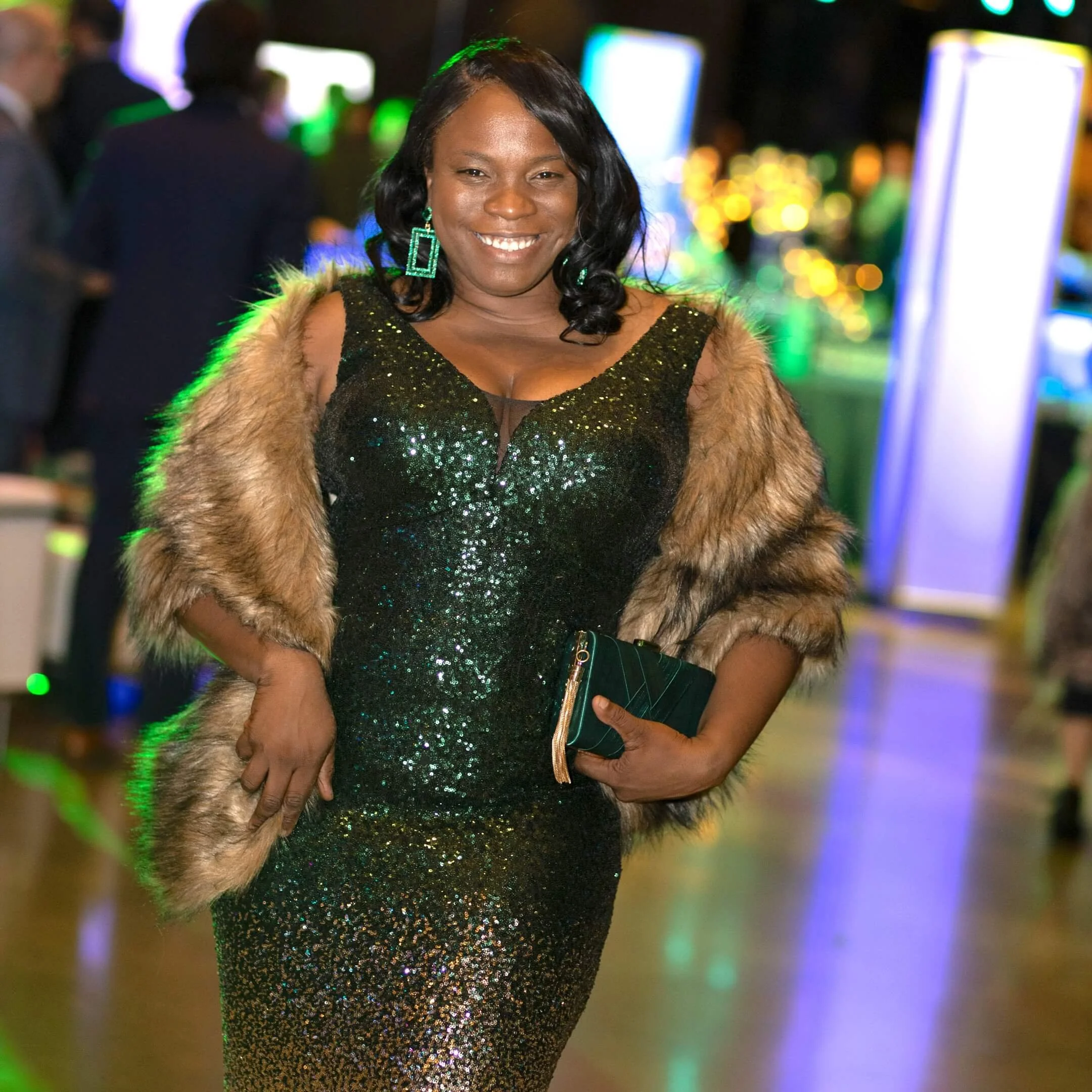 A woman in a black sequin dress holding a clutch purse, wearing a fur stole, and smiling at an event with colorful lights in the background.