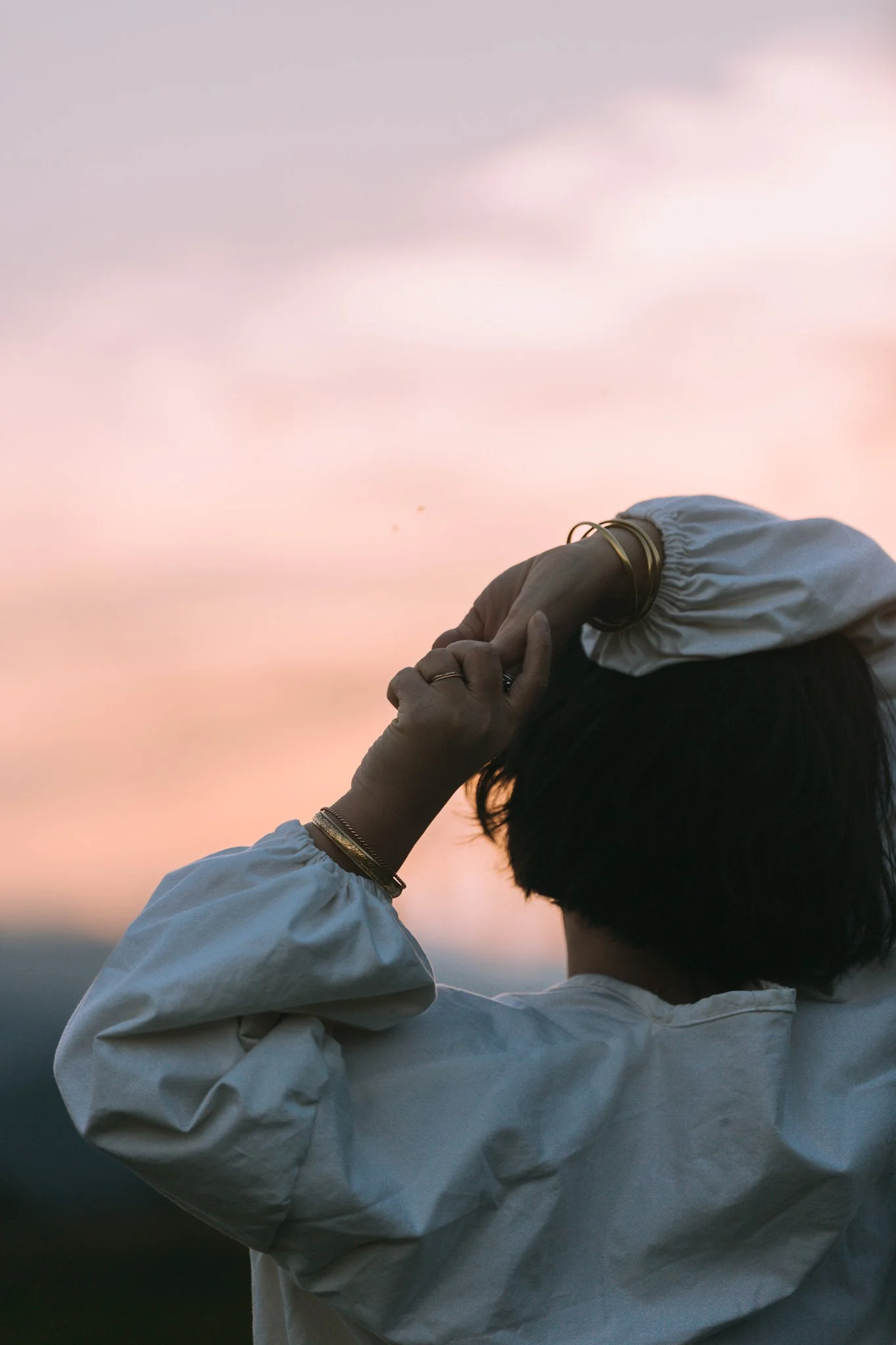 A person with short dark hair, seen from behind, holding their head with both hands against a soft pastel sunset sky, wearing a light-colored jacket multiple bracelets, and rings.