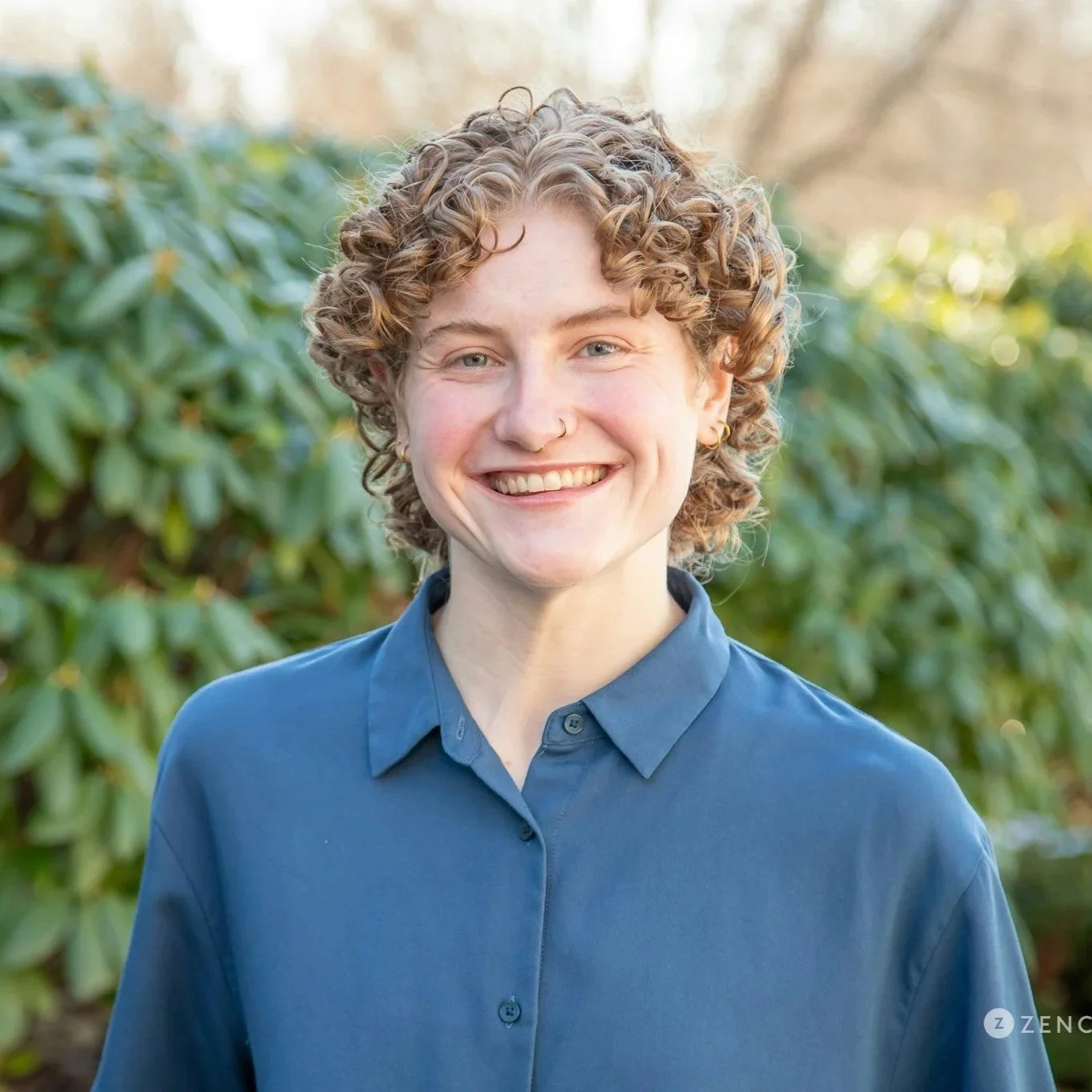 A smiling young person with curly blonde hair and a septum piercing in front of autumn-colored leaves on a tree.
