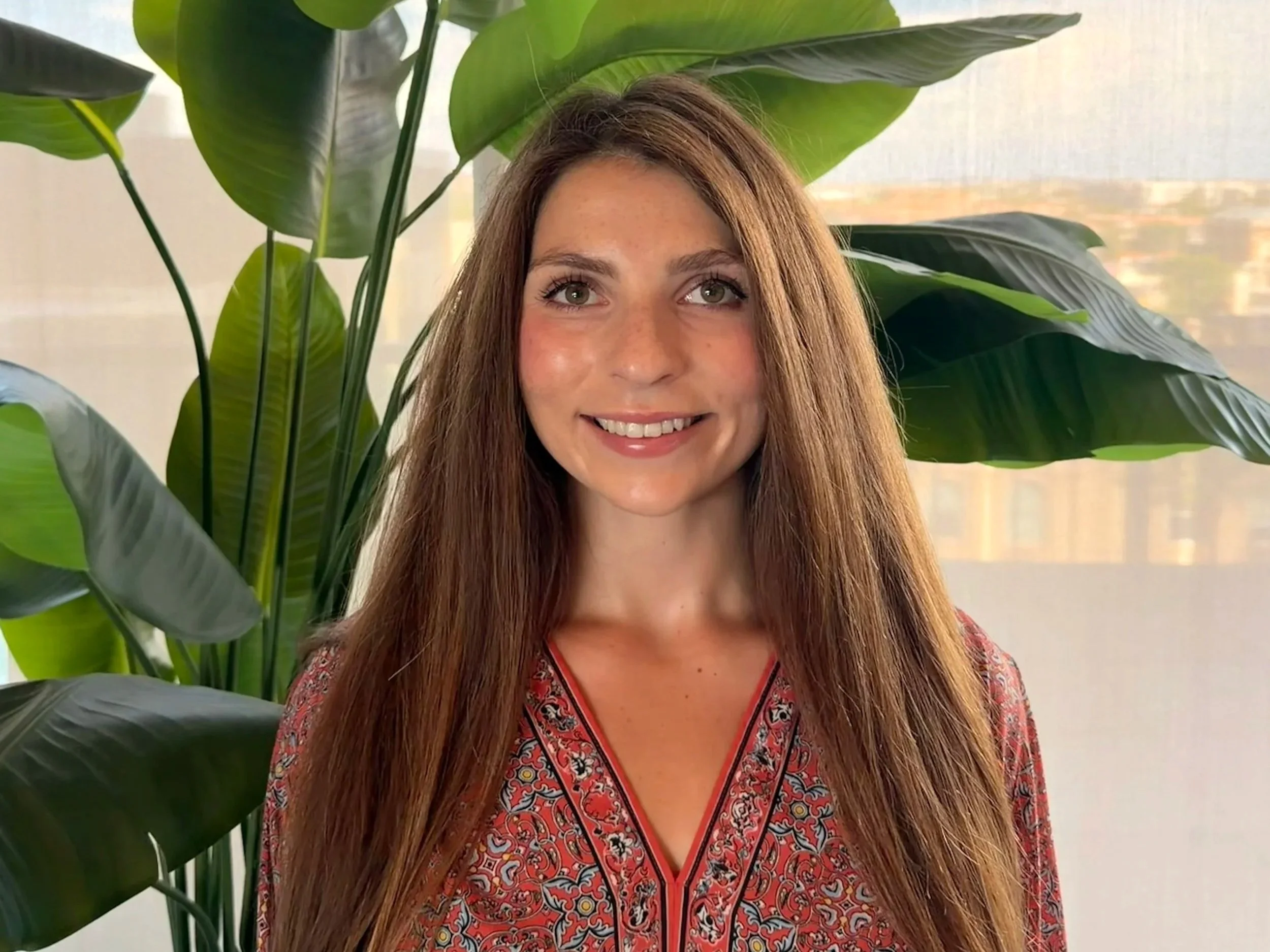 A young woman with long brown hair and blue eyes smiling indoors near a window with a brick wall and green houseplants in the background.