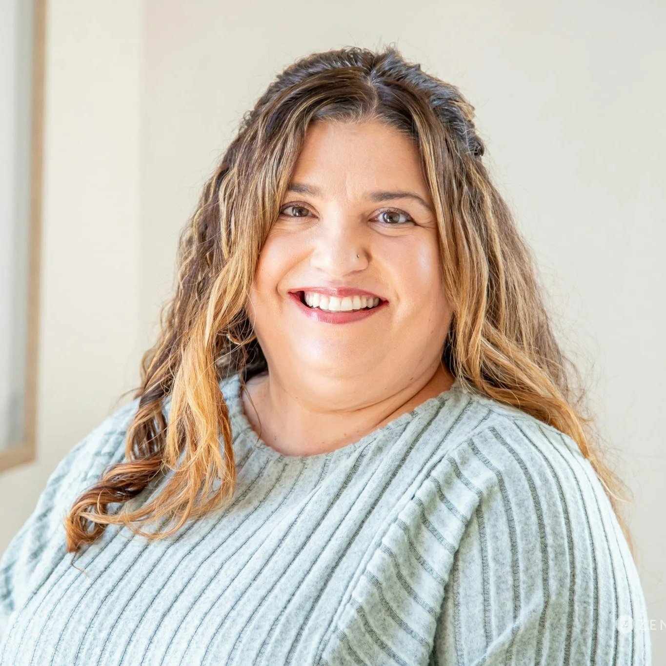 Woman with curly hair smiling, wearing a floral dress, standing amidst lush green plants.