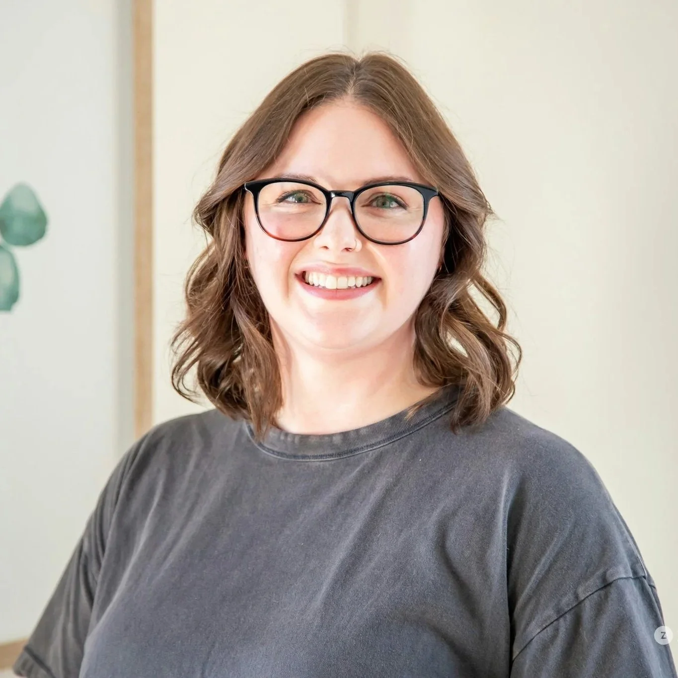A woman with long wavy brown hair and fair skin, smiling and wearing a teal shirt, stands outdoors in front of green foliage.