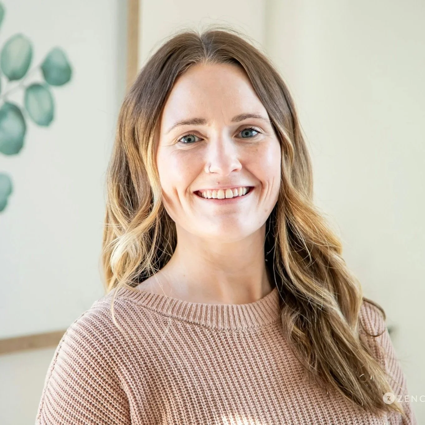 A woman with long, light brown hair, wearing a black textured top, smiling at the camera against a plain beige background.