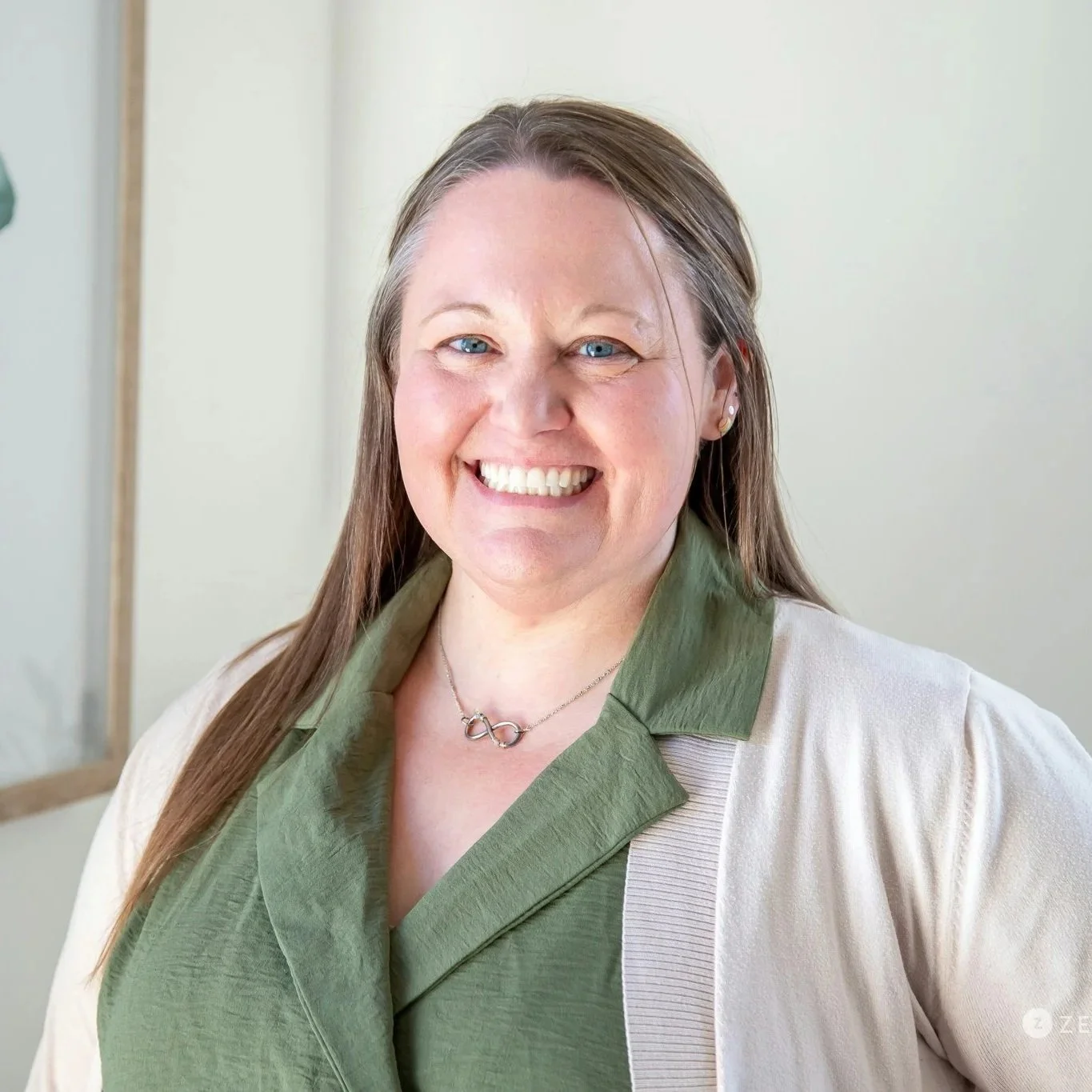 A smiling woman with long hair, wearing a red shirt and black jacket, standing in front of a light-colored wall with framed artwork.