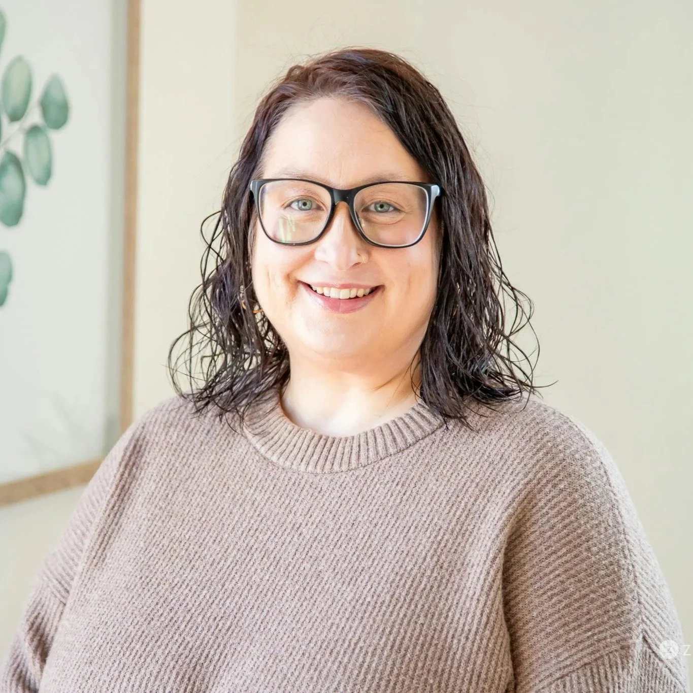 A woman with dark brown wavy hair, wearing glasses and a nose ring, smiling for a selfie on a porch with a blurred background of trees and a house.