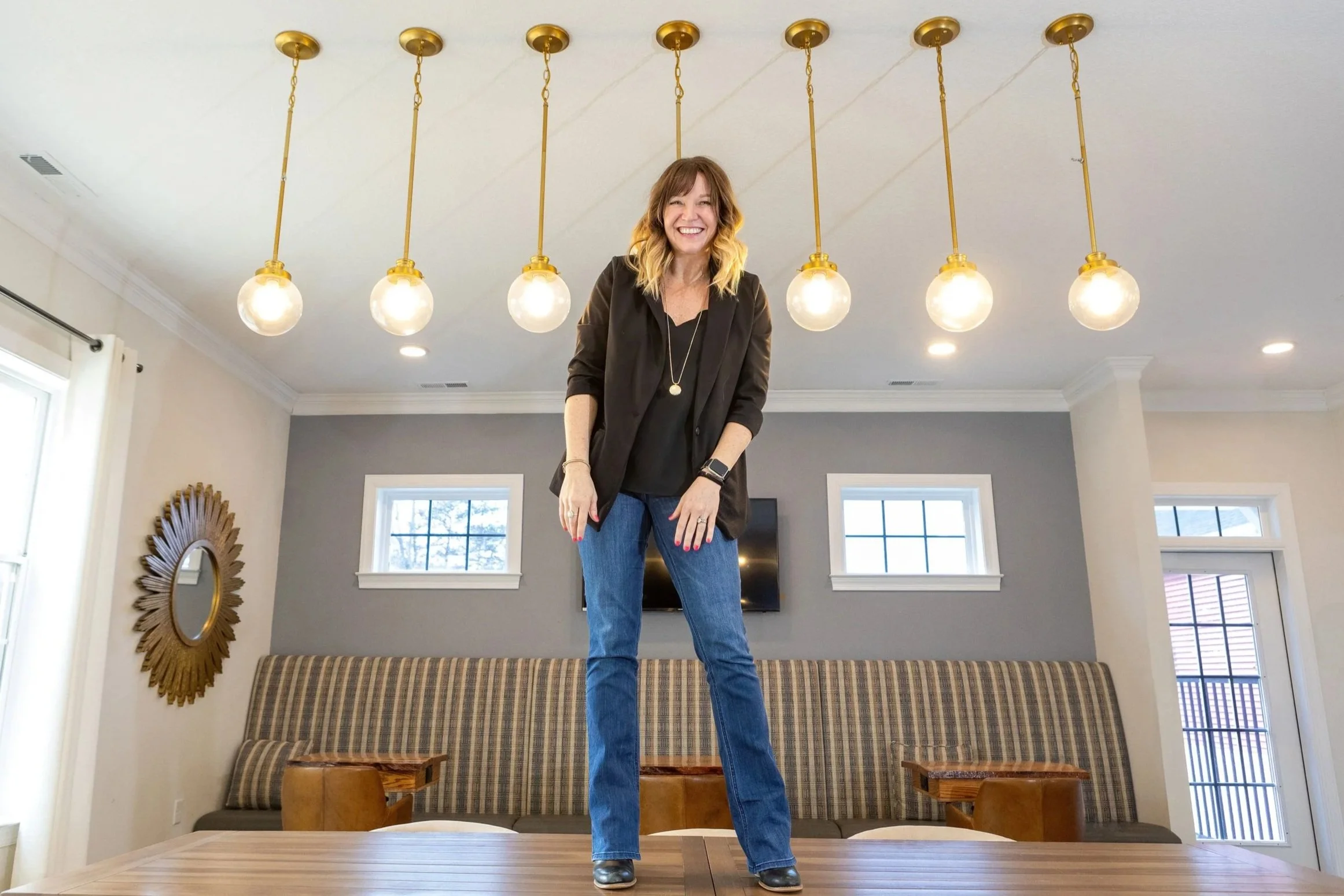 Alyssa Kelley of AWH Interiors standing playfully on a table beneath modern gold pendant lights, embodying her creative, approachable design style.