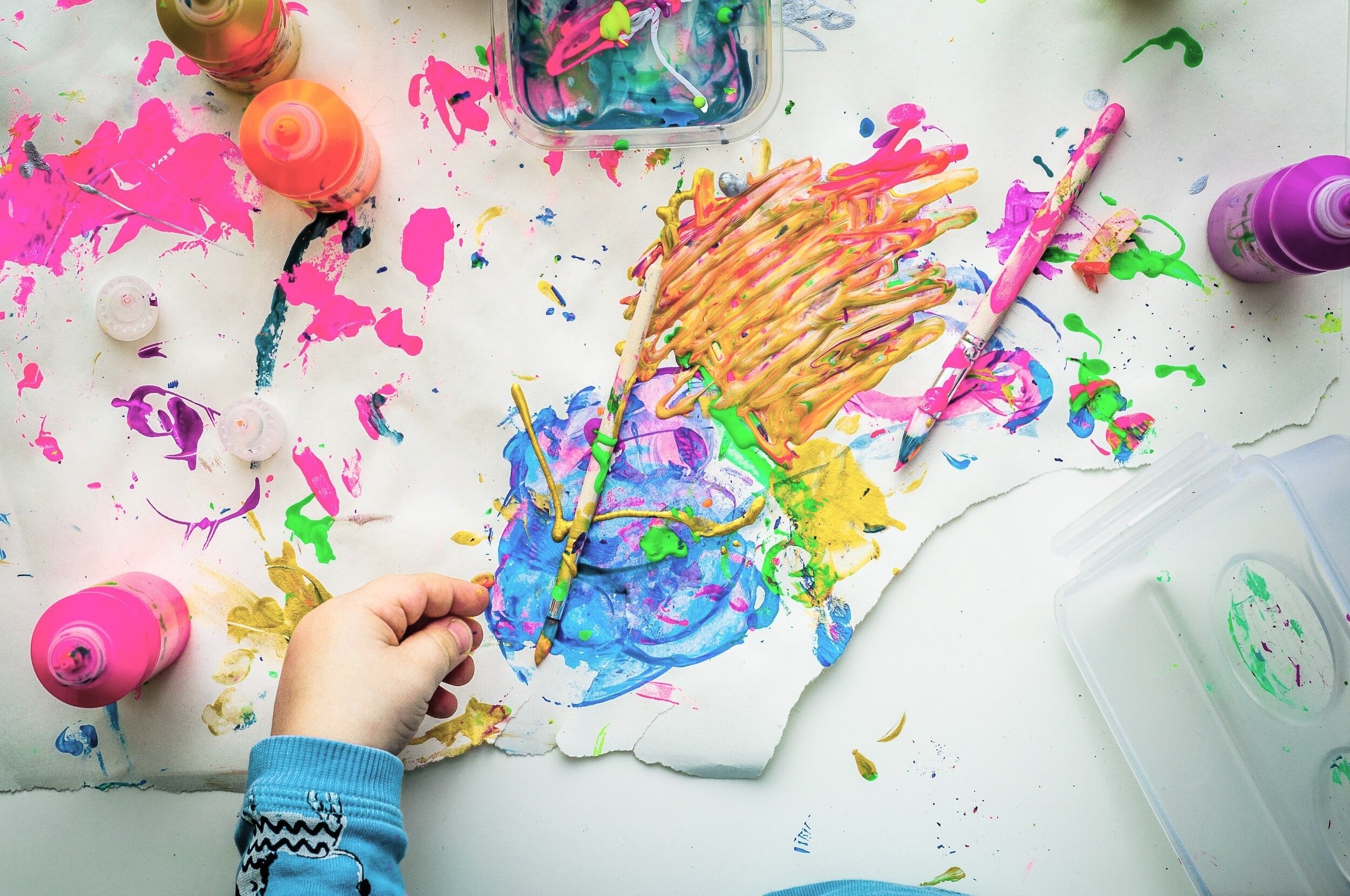 Child's hand holding a paintbrush over a messy table with colorful paint splatters, drips, and blobs on paper and surrounding area, with bottles of bright paint and paint containers scattered around.