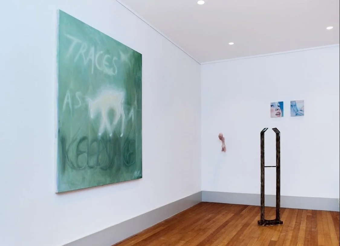 An art gallery with white walls and wooden floors. Large abstract painting with green background and white text on the left, two small square photos of a woman's face and a hand on the right, a small sculpture of a human face and a wooden sculpture i