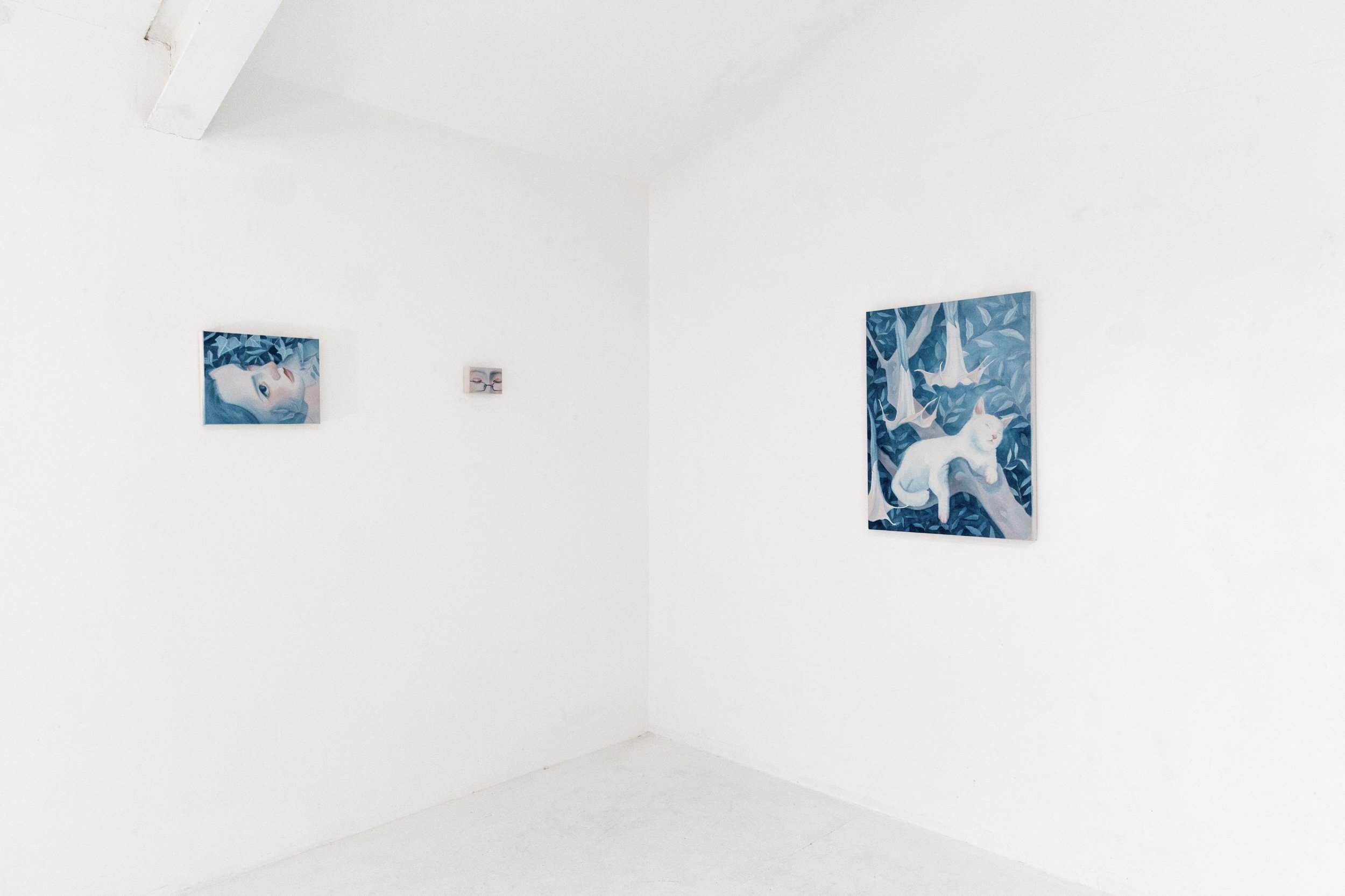 Art gallery wall with three framed paintings: one on the left depicting a woman's face with pale skin, light hair, and blue eyes; a smaller one in the center showing two glasses of a drink; and a larger one on the right illustrating a white animal, p