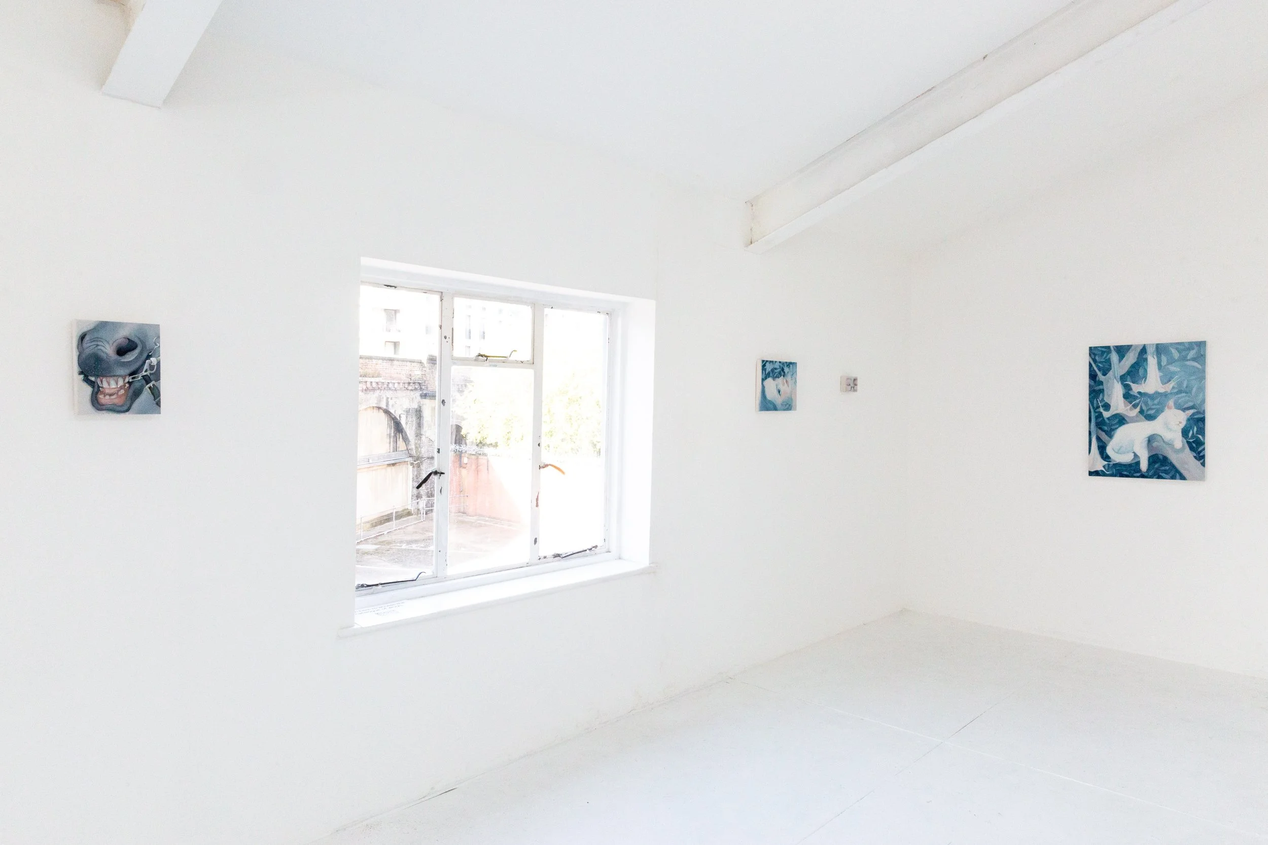 A minimalist white art gallery with three paintings on the walls and a large window showing an urban outdoor scene.