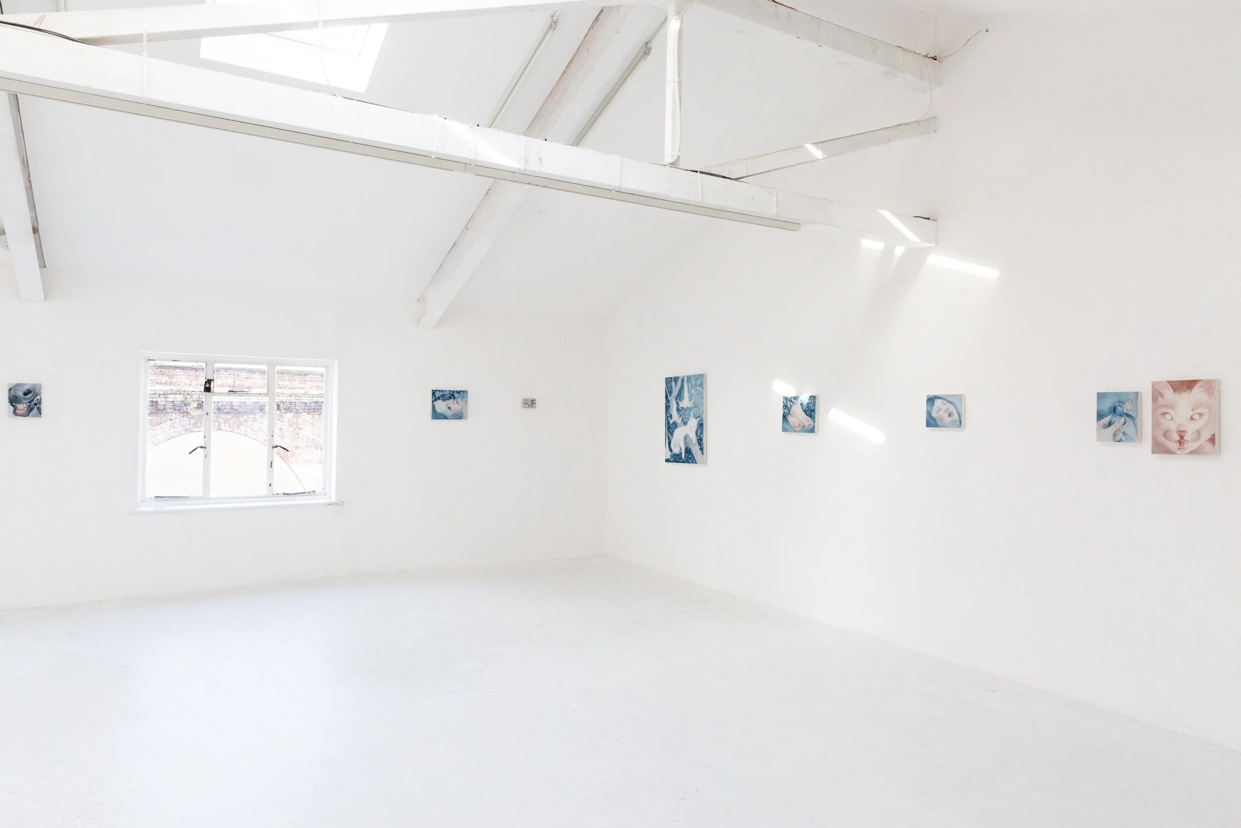 White gallery or art exhibit space with paintings on the walls, a high ceiling with exposed beams, and a window allowing natural light into the room.