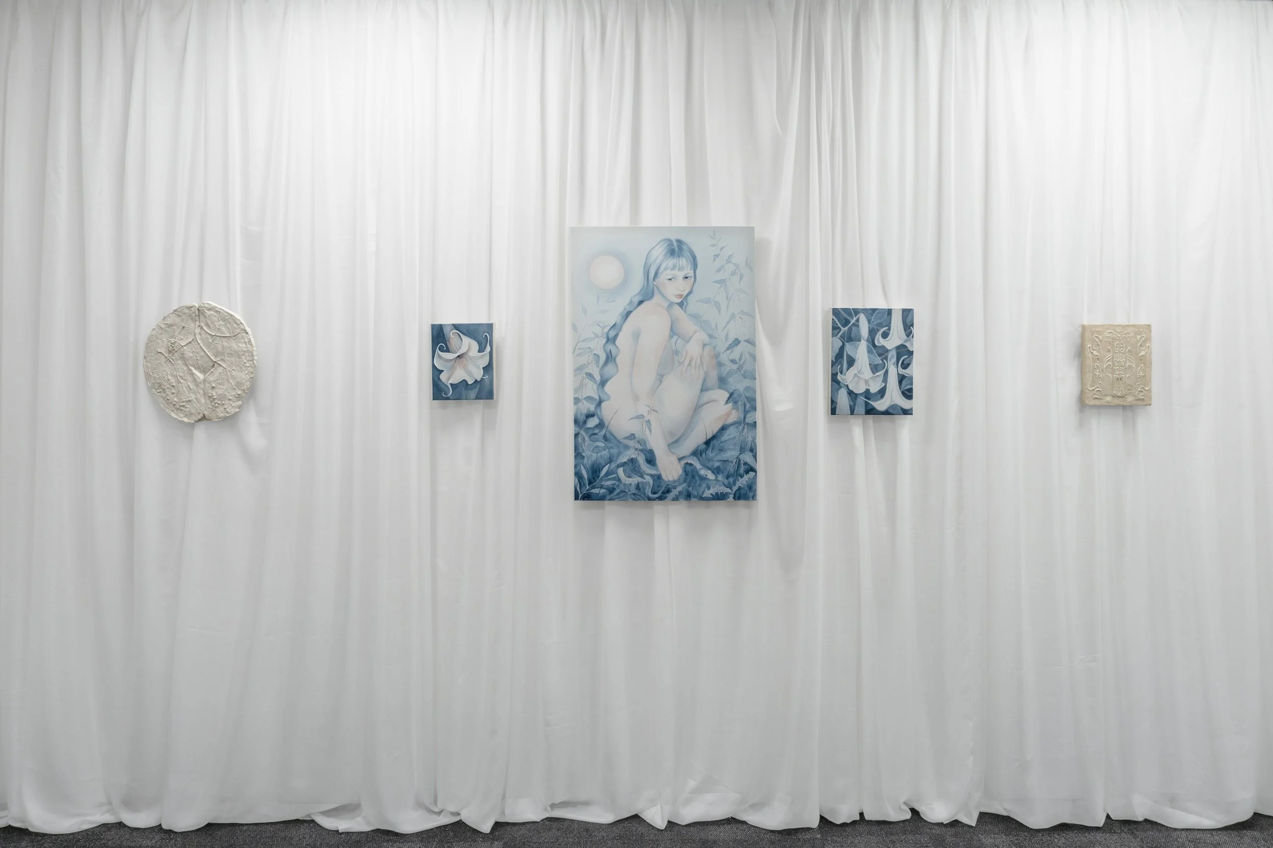 An art display with five artworks mounted on a white fabric backdrop. The central piece is a large portrait of a woman with long, wavy hair, sitting amid foliage under a moonlit sky. To its left and right are smaller, similar art pieces featuring abs