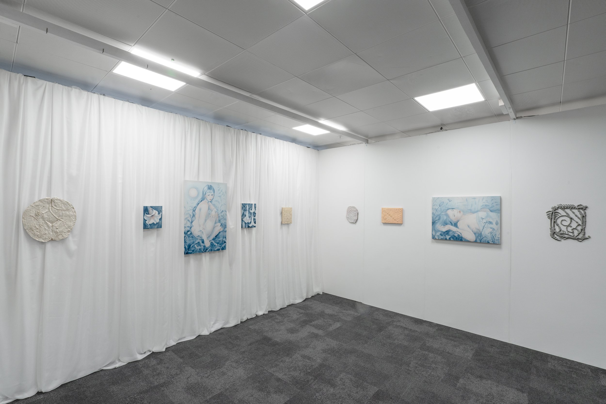 Art gallery with white curtains and blue-themed artwork, including paintings of women, textured wall art, and metal sculptures, under fluorescent ceiling lights.