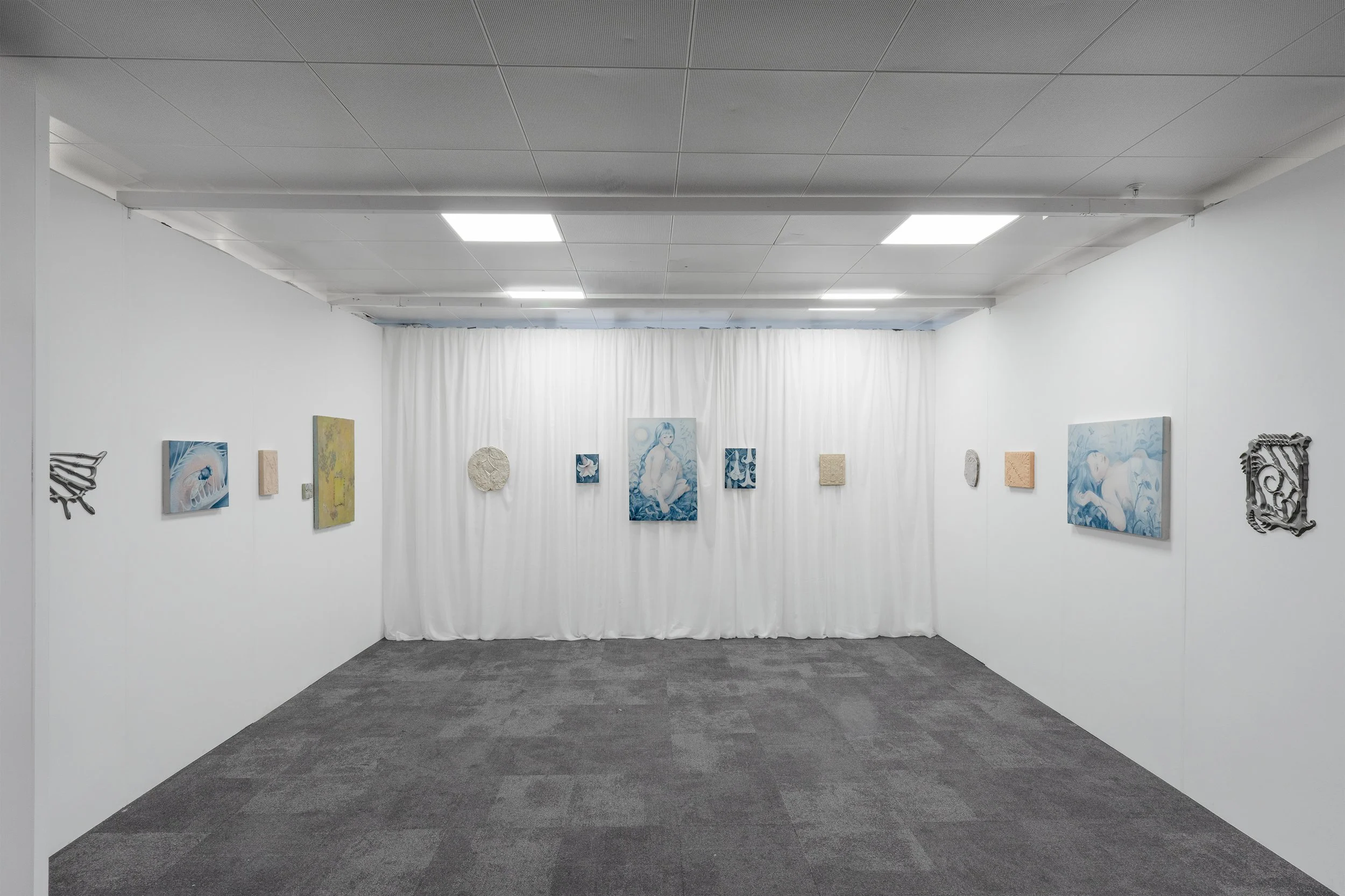 An art gallery with white walls and a dark gray carpeted floor, displaying various paintings and sculptures. The back wall is partially covered by a white curtain, with artworks hanging on it.