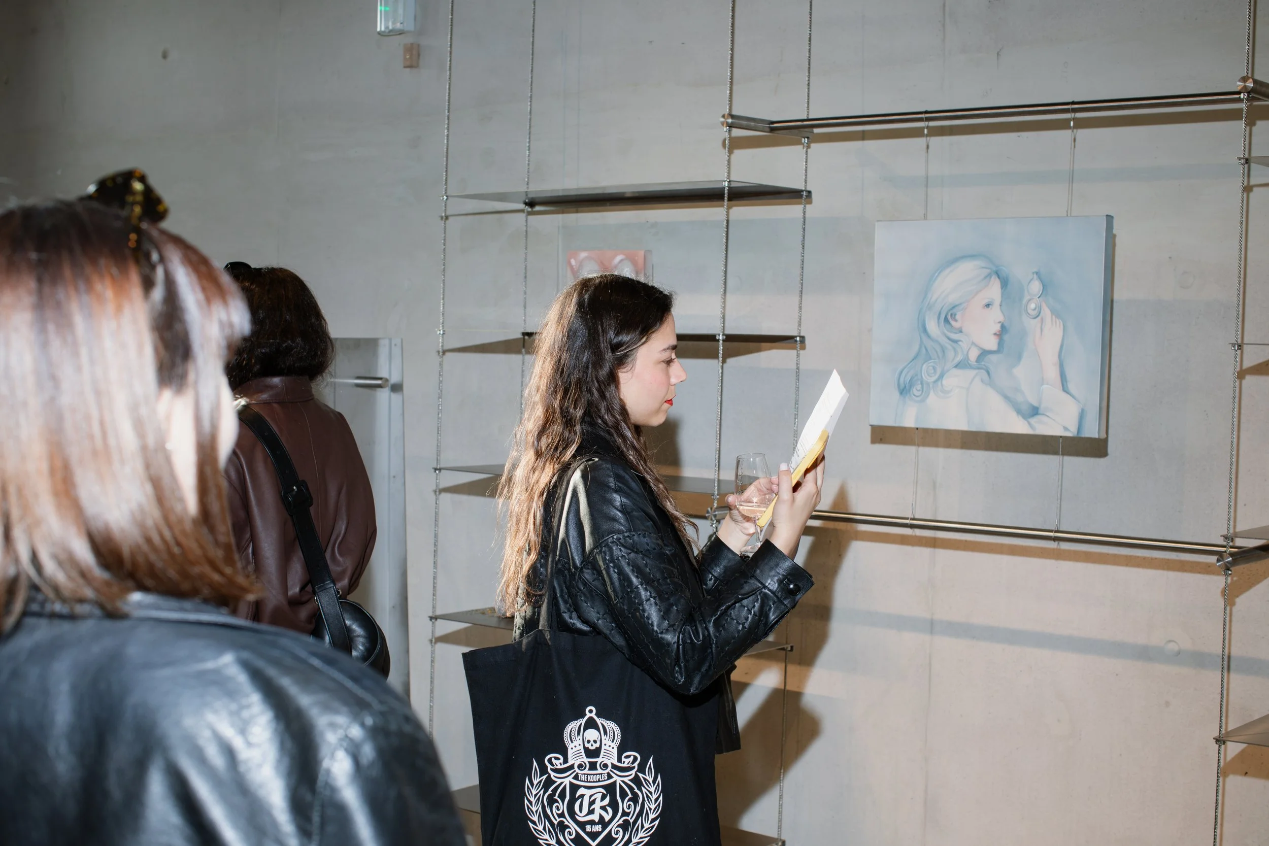 A woman with long dark hair reading a book or menu at an art gallery with others watching. There is a painting of a woman with long hair holding a small object on the wall behind her.