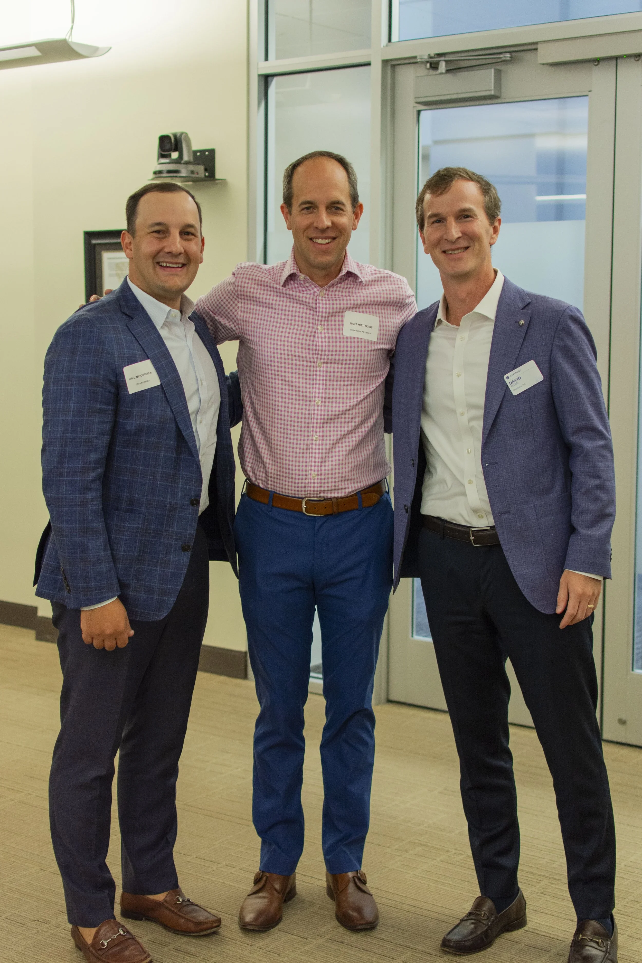 Three men in business attire smiling and standing together indoors, with a woman in the background, near glass doors and windows.