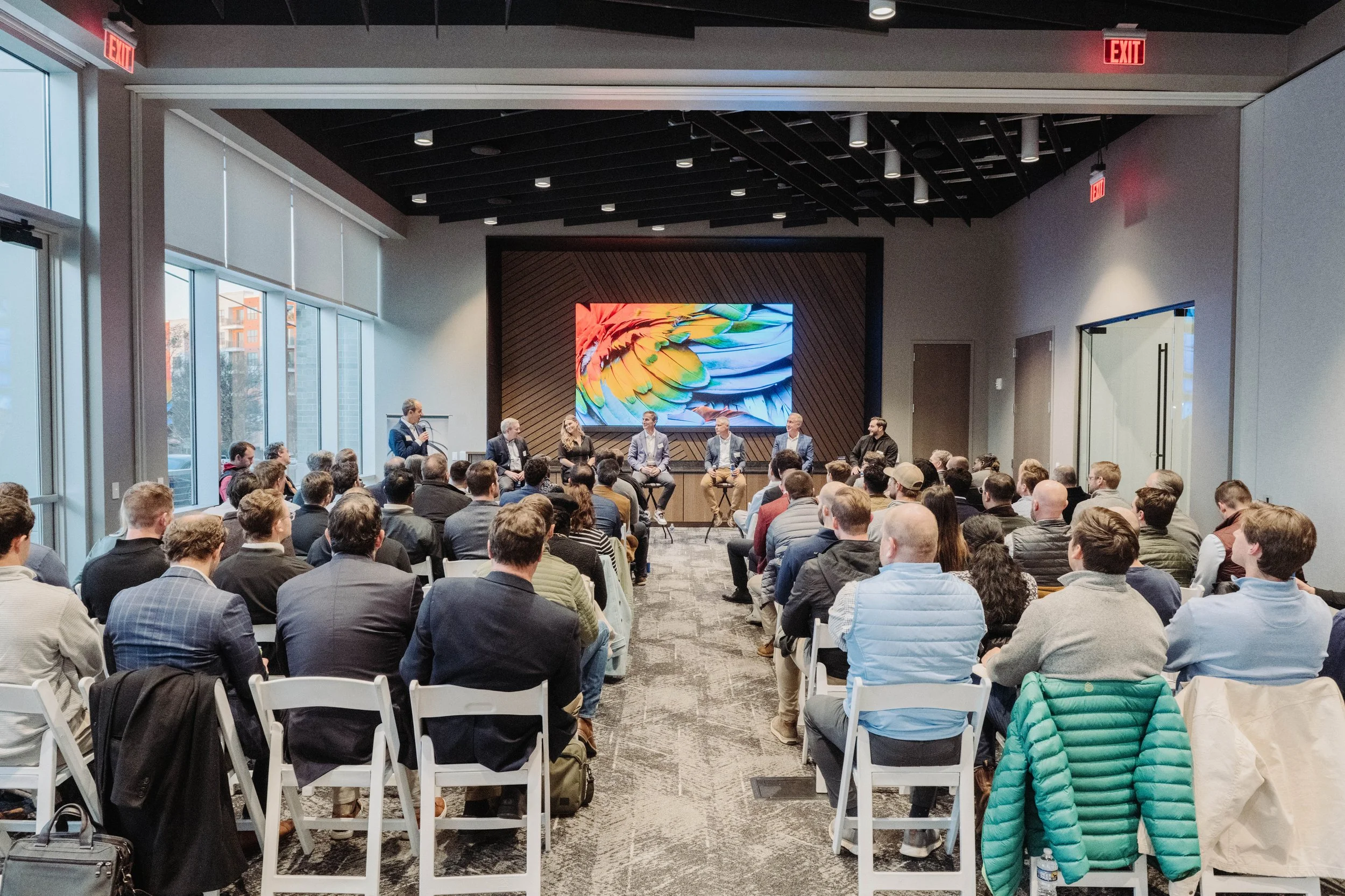 A panel discussion or presentation event taking place in a modern conference room with a large audience. Six panelists are seated on stage in front of a colorful digital display, with one person standing and speaking into a microphone. The room has large windows on the left and modern architectural features.