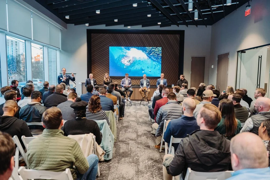 A conference room filled with people attending a panel discussion or presentation. There are six panelists seated at the front, with one person standing and speaking to the audience. Large windows reveal an urban cityscape outside.