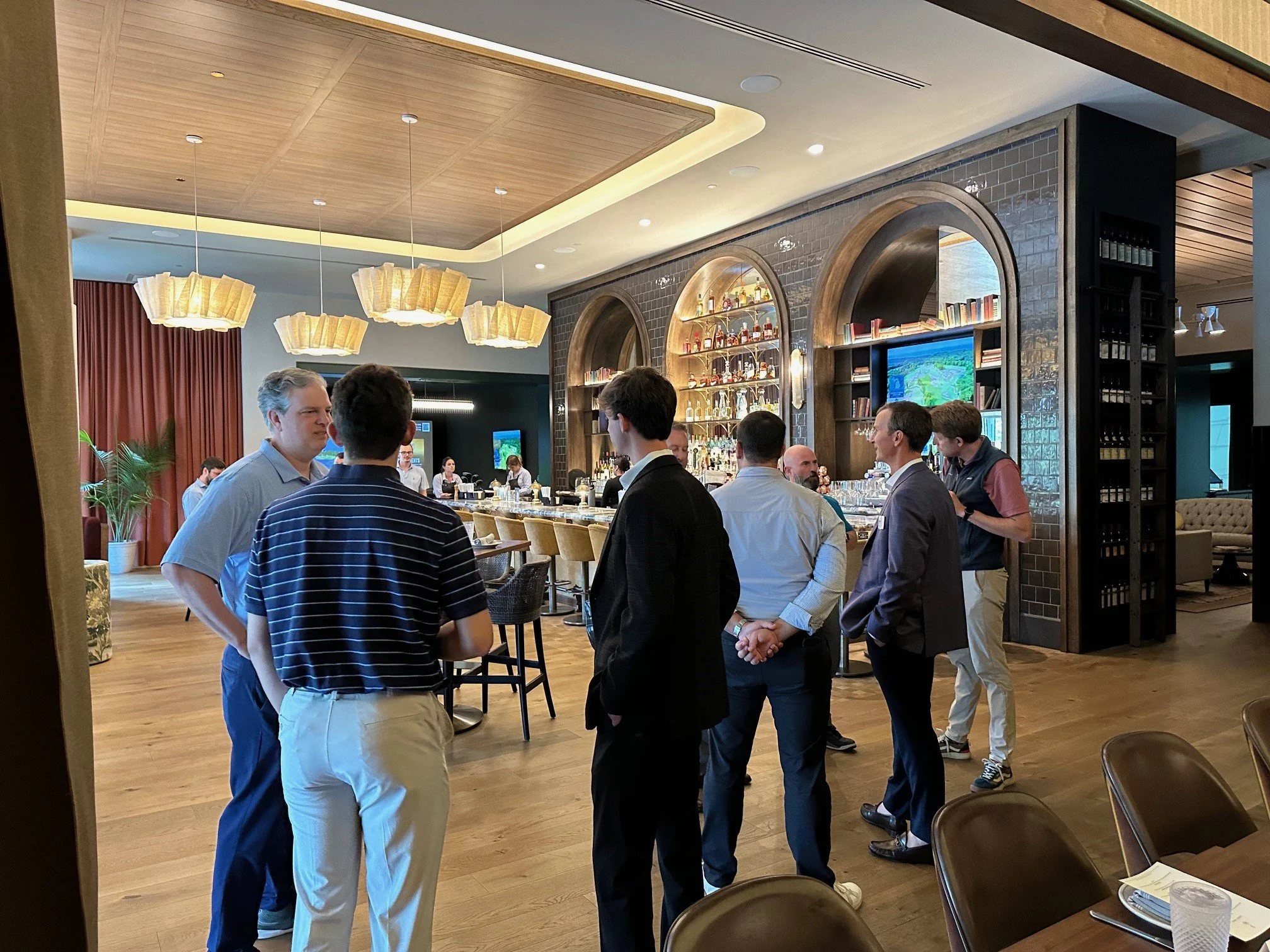 A group of people gathered and talking inside a modern restaurant or bar with wooden and brick interior decor, and hanging light fixtures.