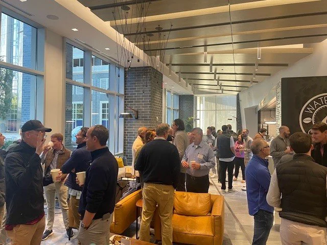 People gathered in a modern, well-lit indoor space with large windows, a brick pillar, and contemporary decor, socializing and holding drinks.