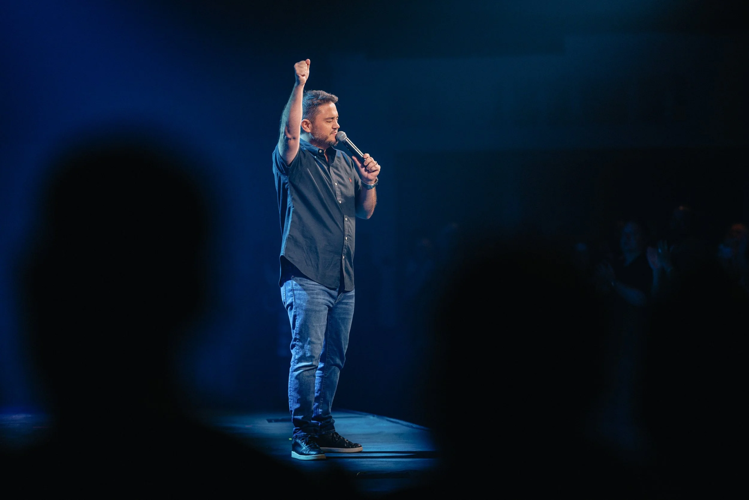 Man giving a speech or performing on stage with a microphone, raising his fist, in front of audience with visible applause.