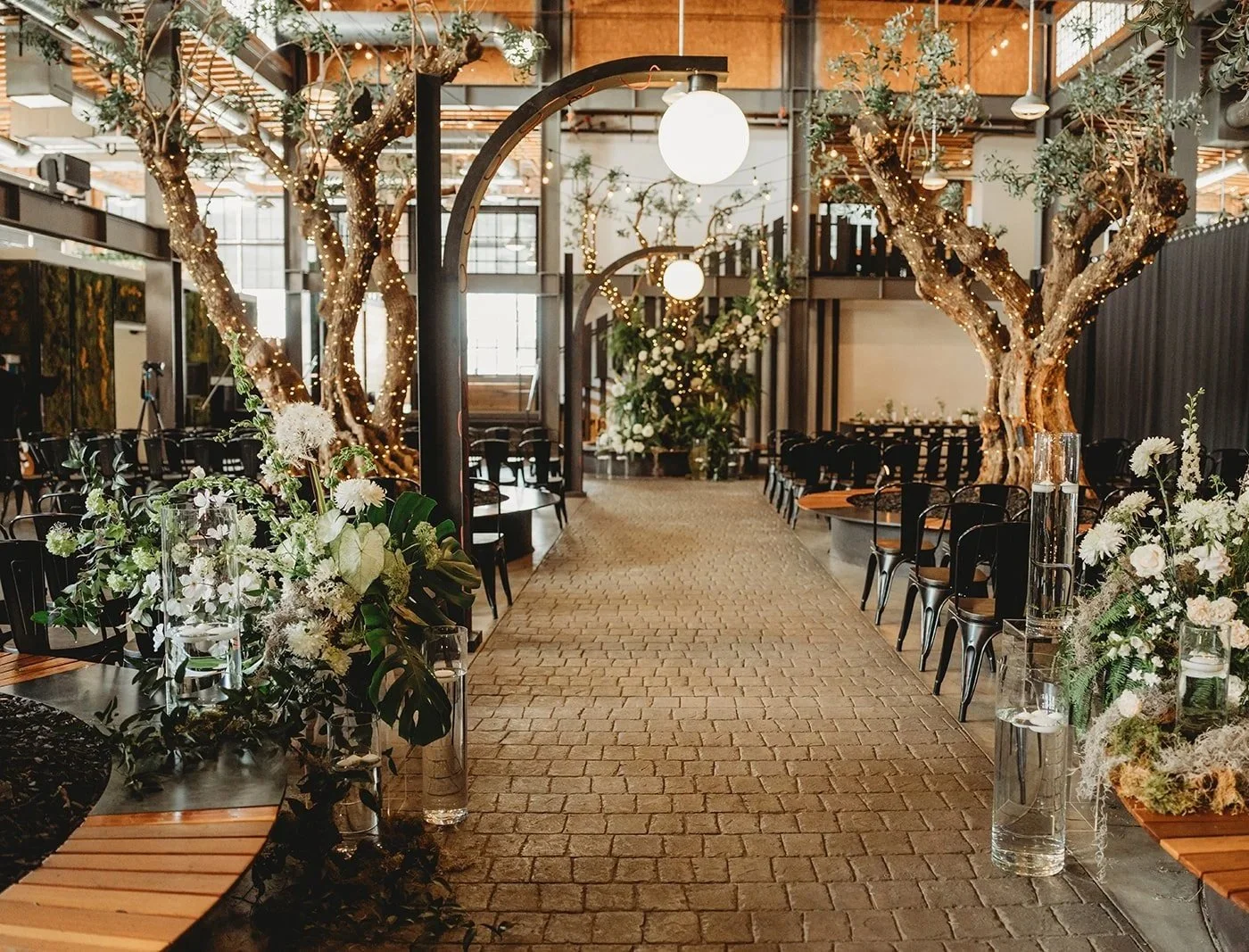 Indoor event space with an olive tree grove (Located at Venue 808). The space has white tables and chairs, with warm lighting and a rustic, modern design.